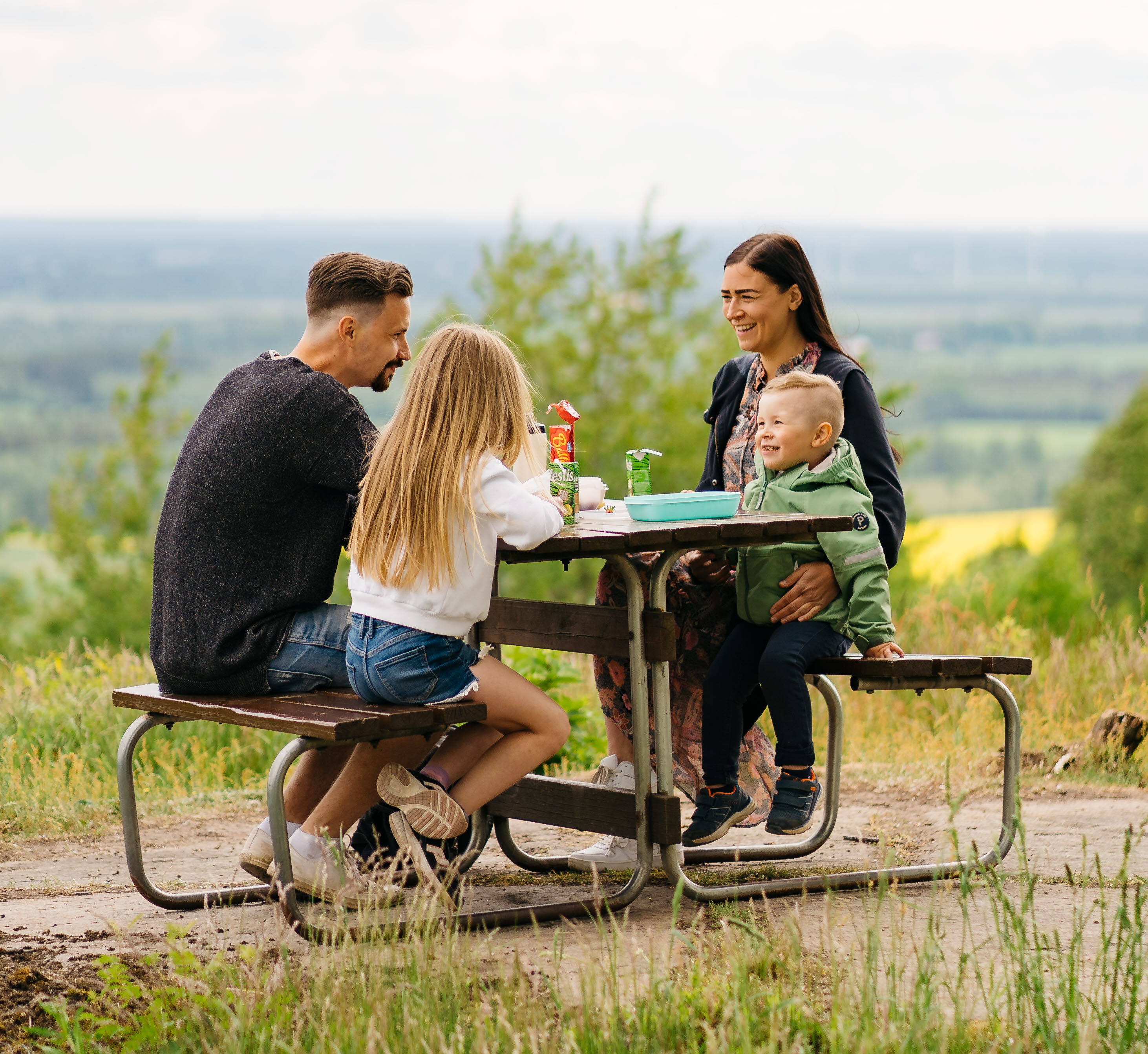 Familj, man, kvinna och två barn sitter och fikar vid bord och bänkar.