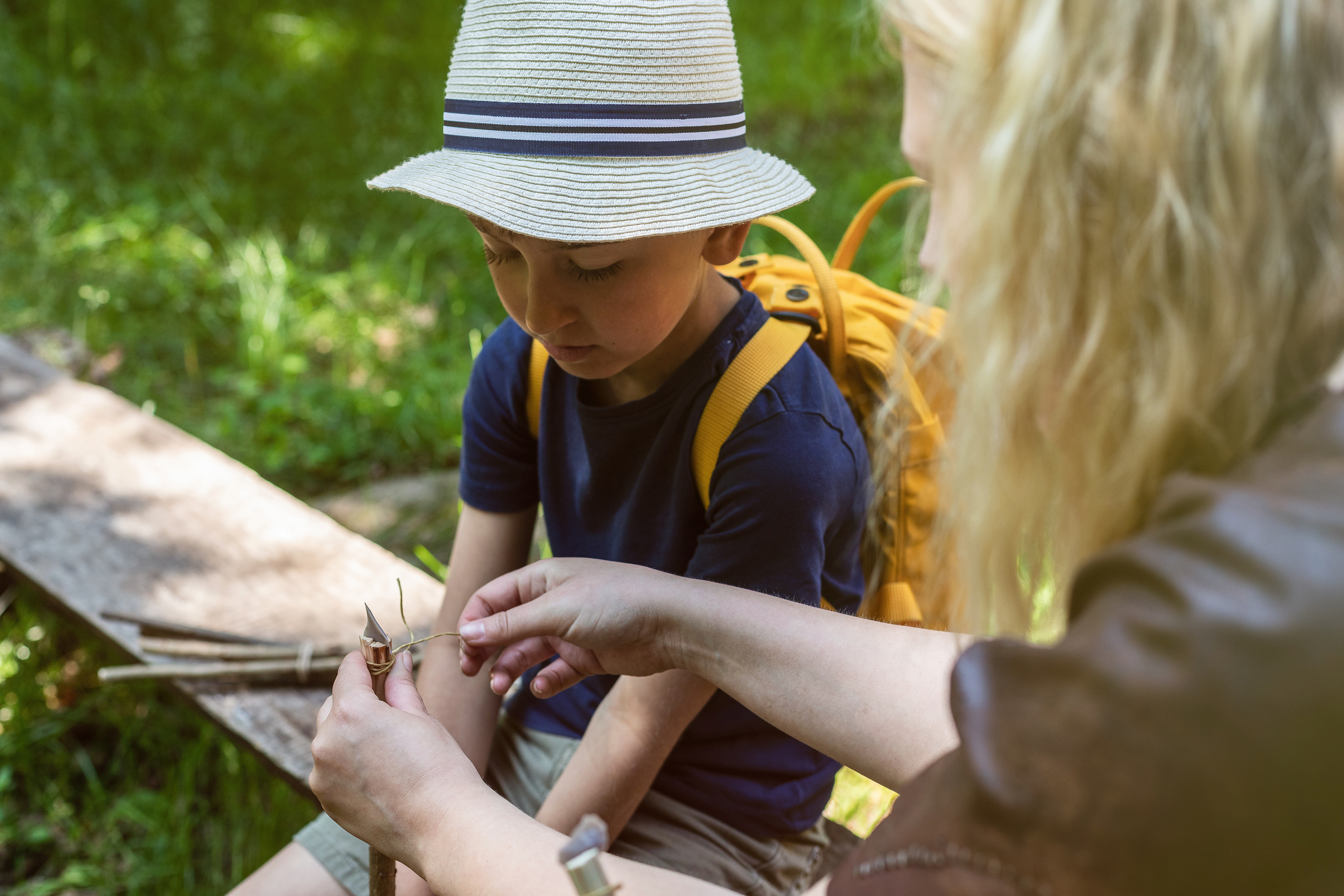 Pojke med hatt som sitter och ser när en kvinna knyter ett band runt en pilspets och en pinne.