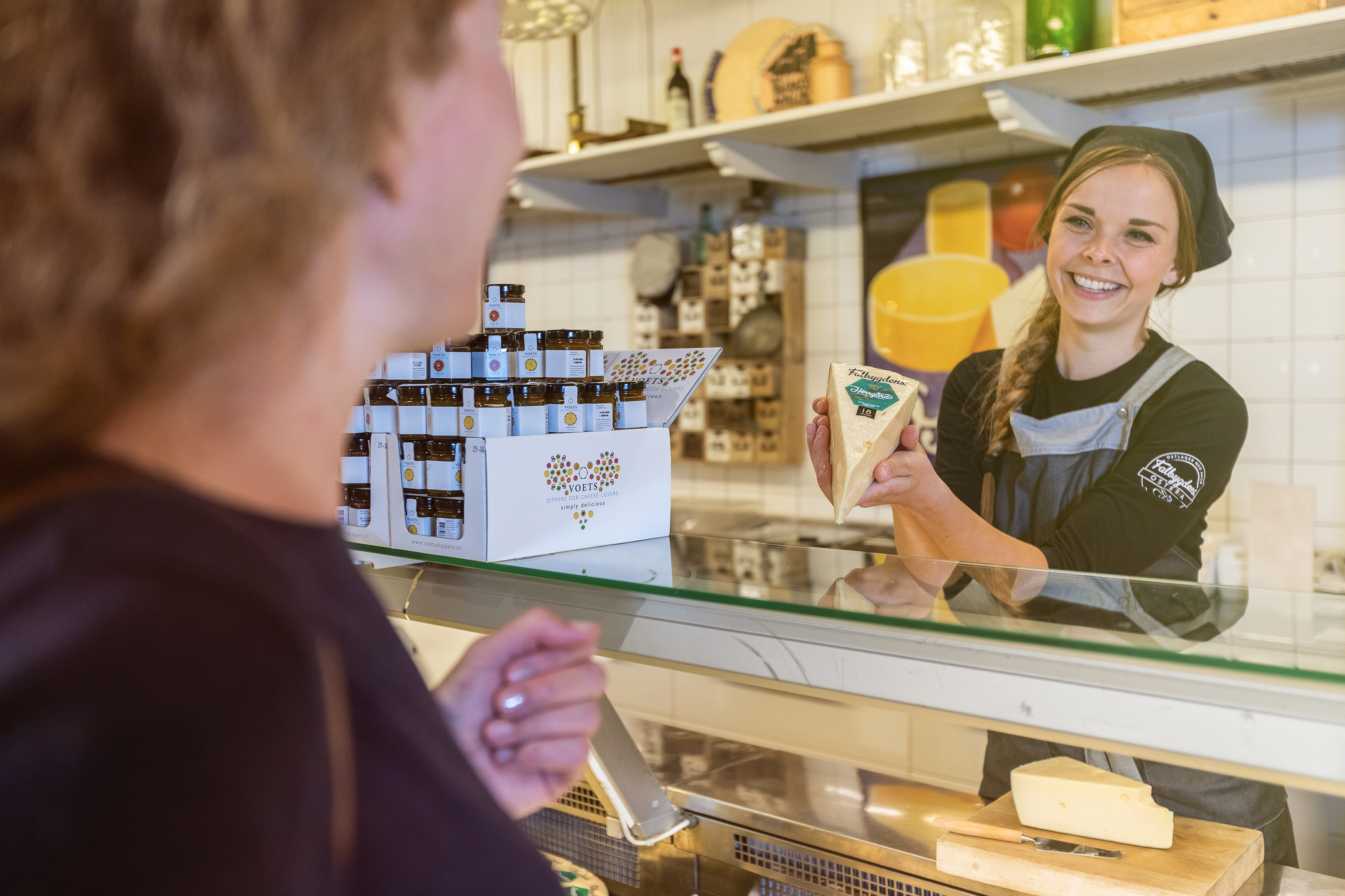Kvinna som står bakom en ostdisk och håller upp en ost för en kund.
