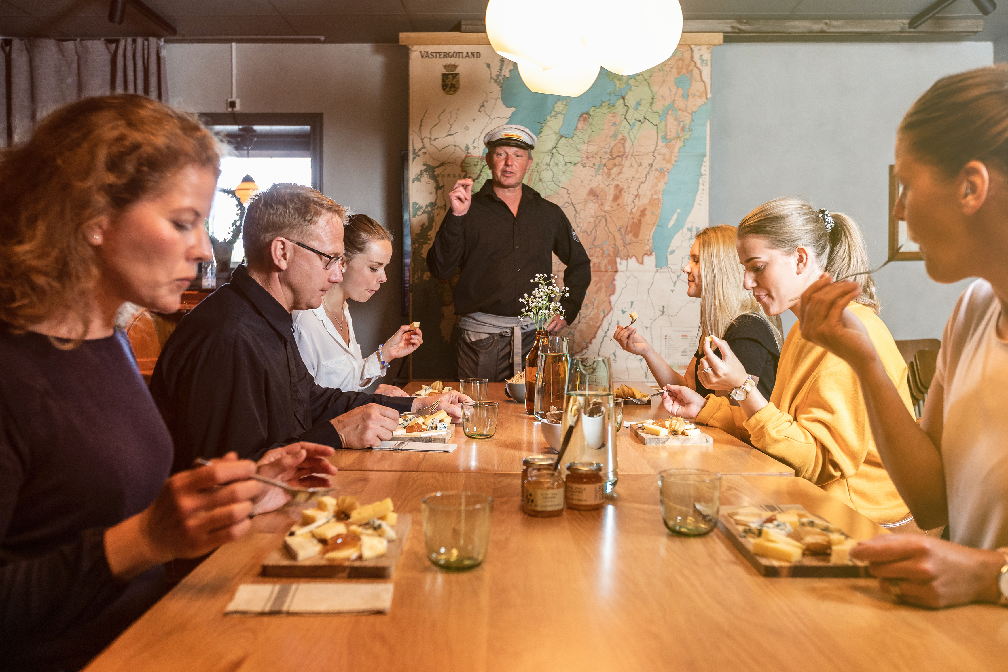 fem personer som sitter vid ett bord med ostbrickor framför sig. en man står och bersättar om ostarna vid kortändan av bordet.