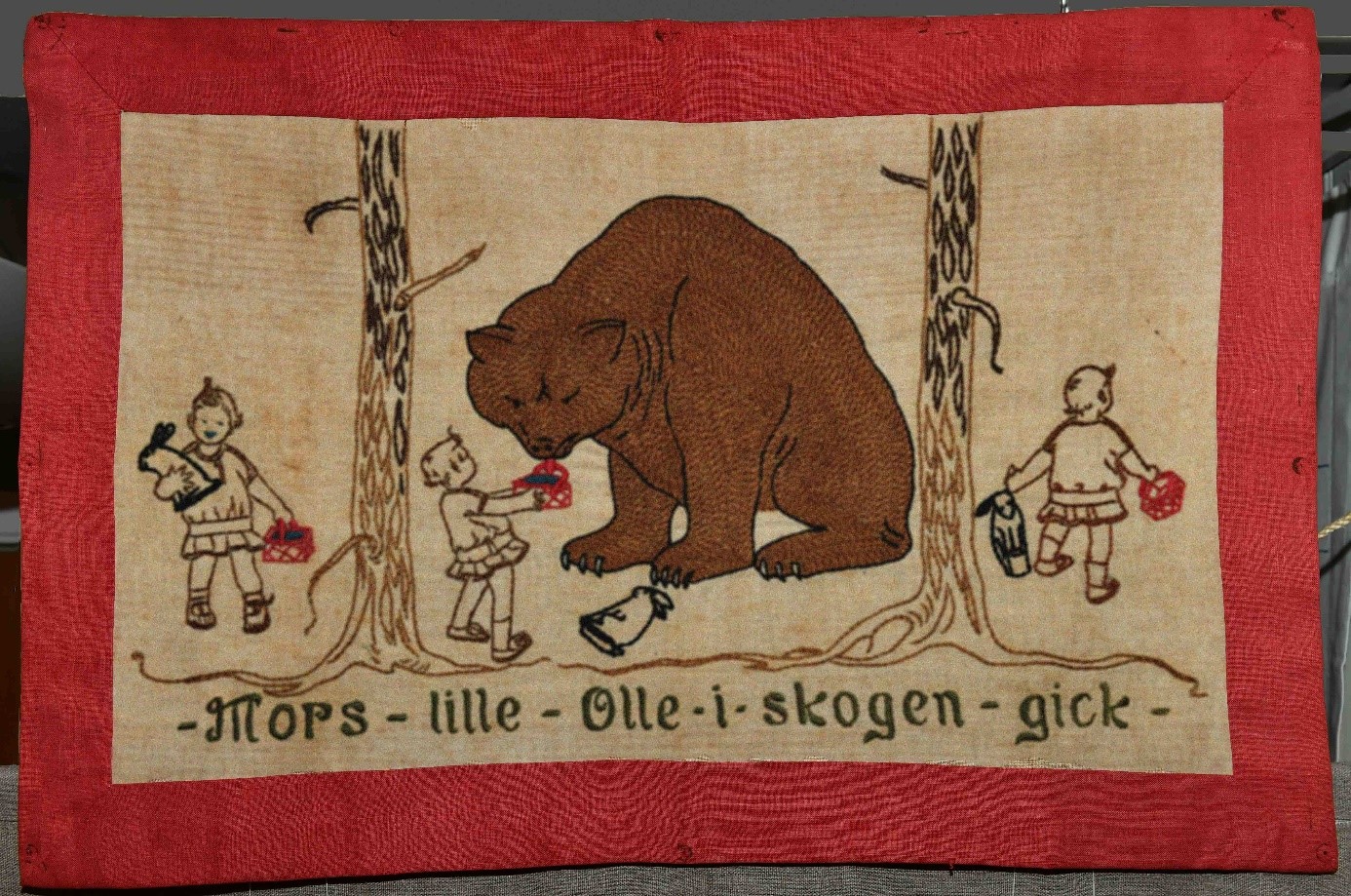 Bonad med en broderad björn och  barn som går i skogen. Texten Mors lille Olle i skogen gick är broderad längst ned.