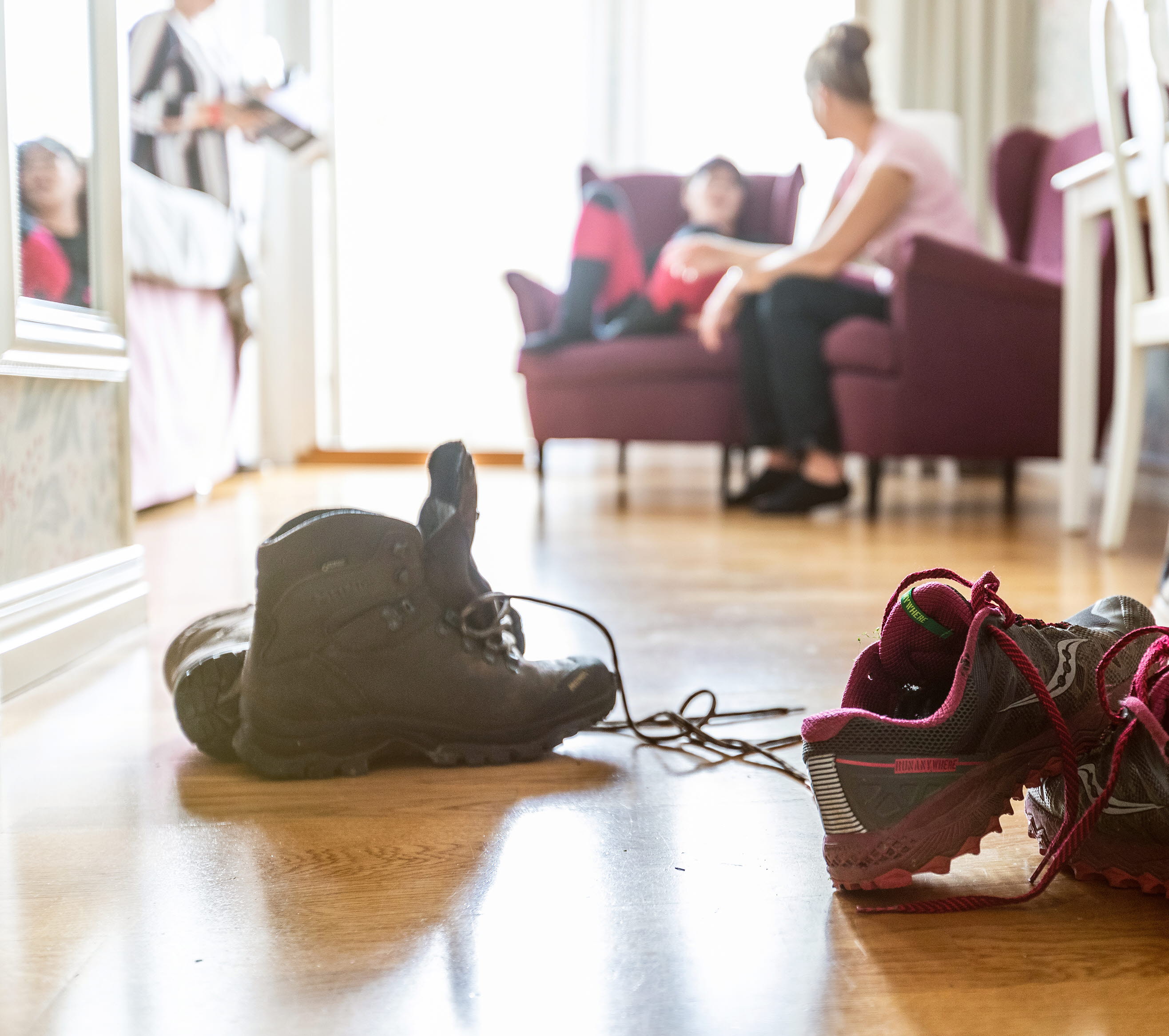 Två vandringsskor står på golvet och två personer sitter i fåtöljer i bakgrunden.