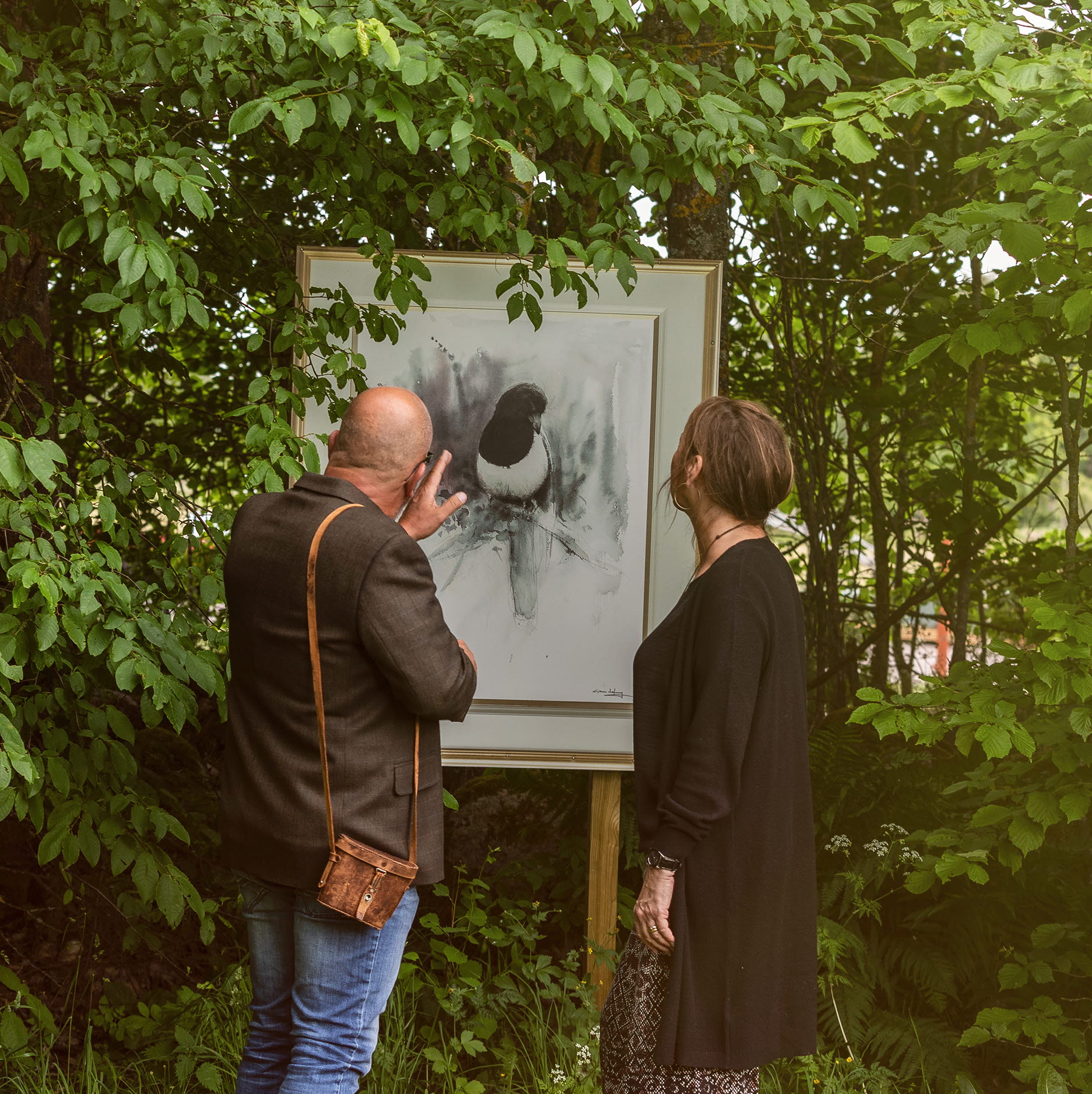 Man och kvinna som kikar på en tavla utomhus bland grönskande blad.