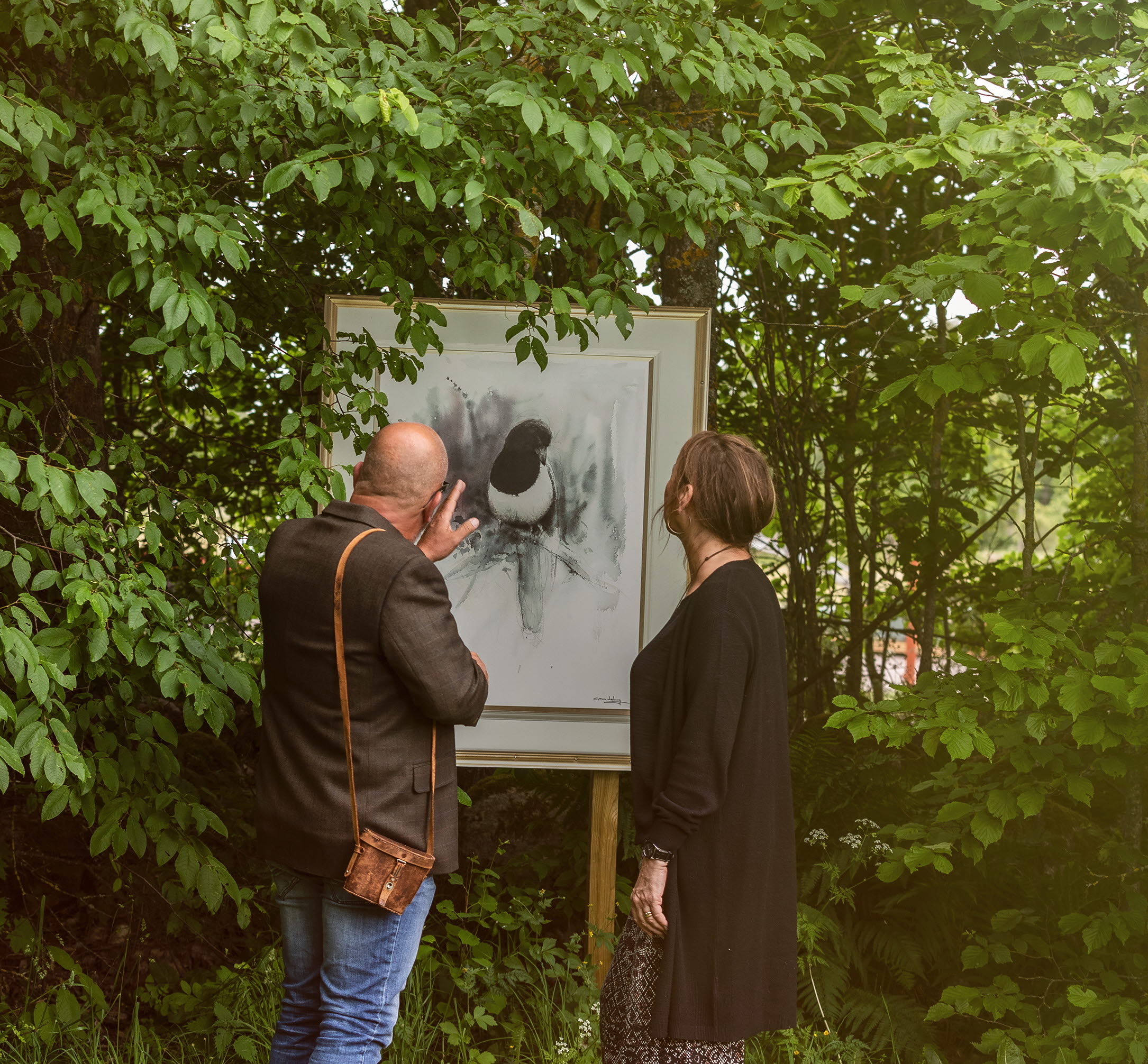 Man och kvinna som kikar på en tavla utomhus bland grönskande blad.