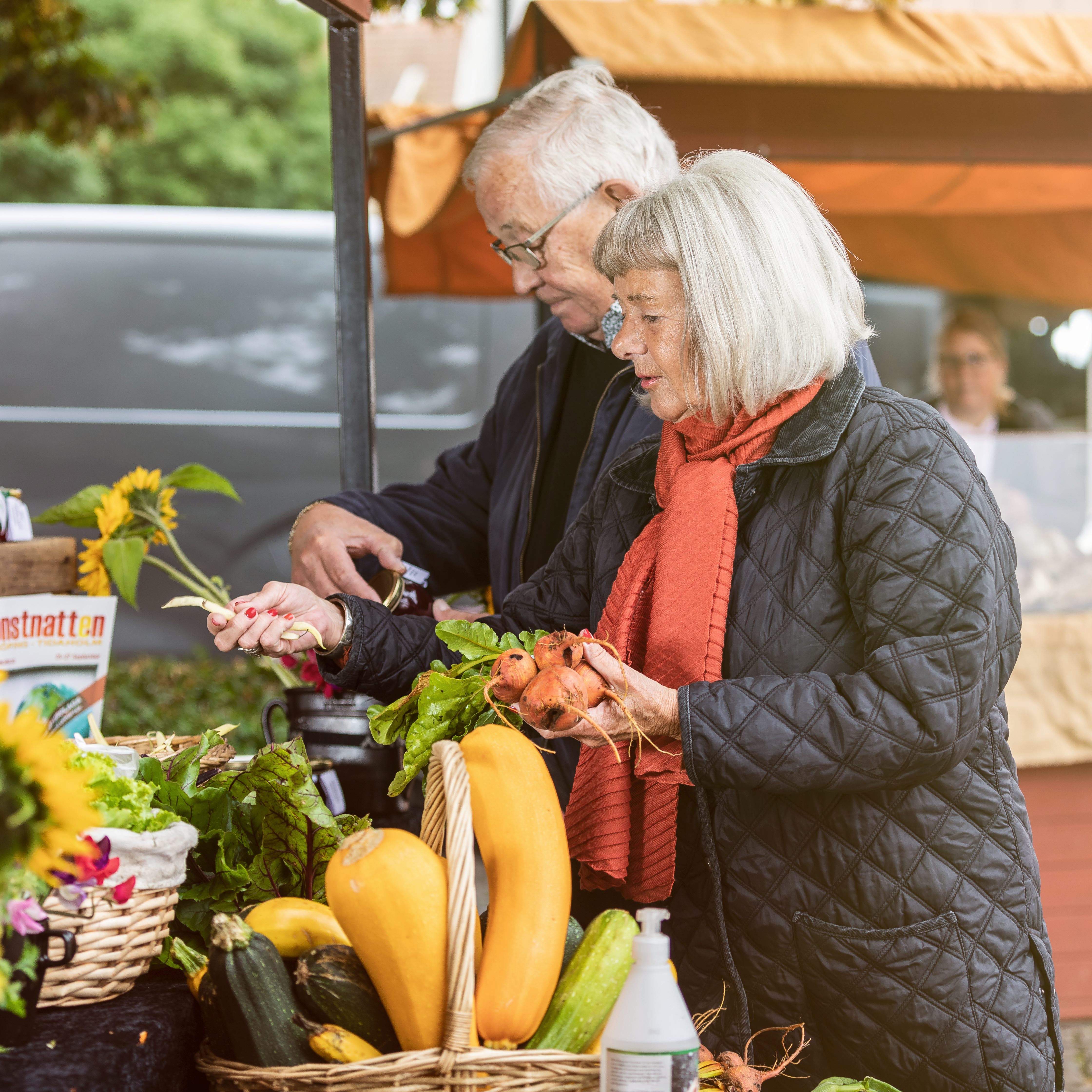 Man och kvinna som står och kikar på olika grönsaker på en marknad utomhus.