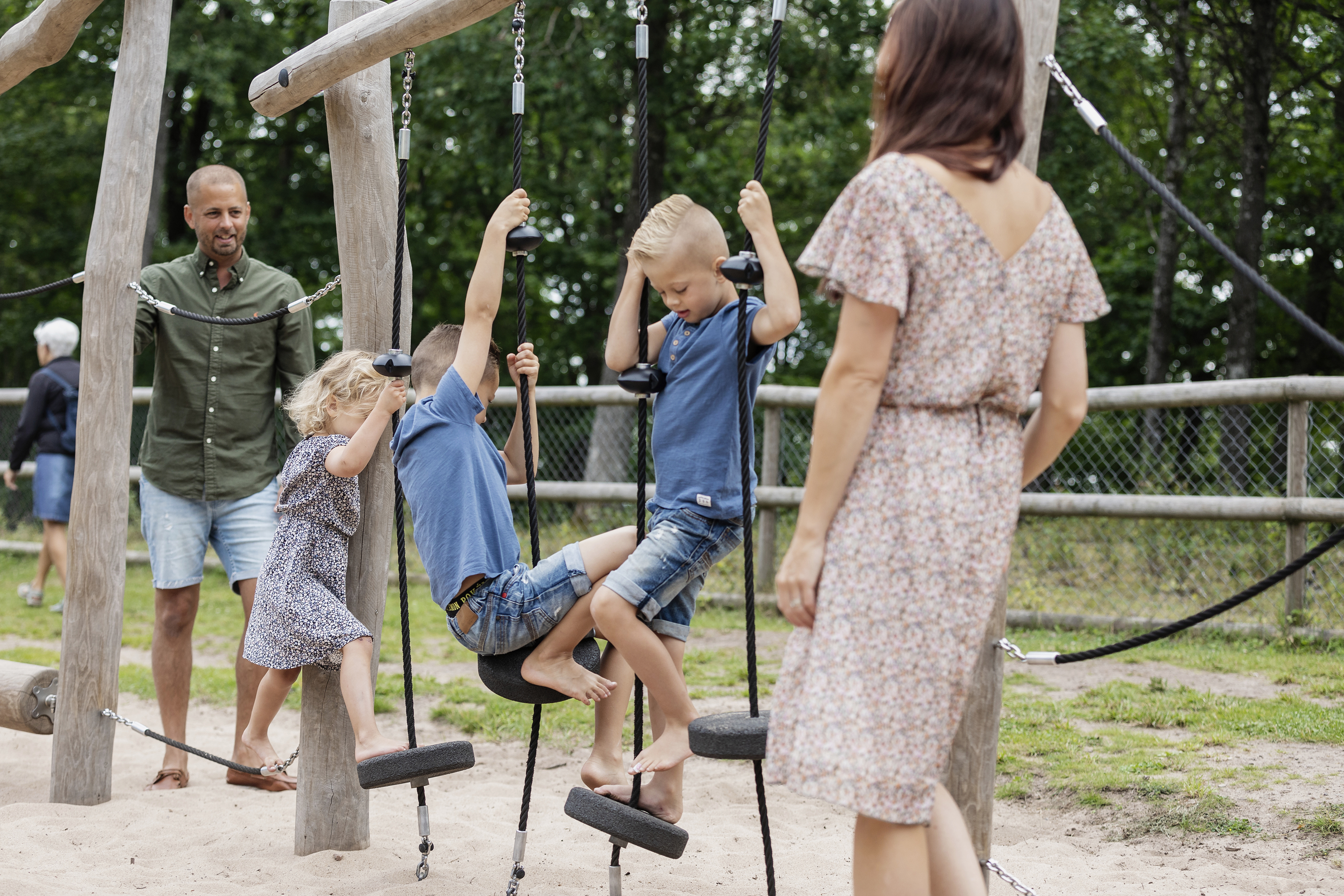 Två pojkar och en flicka klättrar i en klätterställning, föräldrarna ser på.