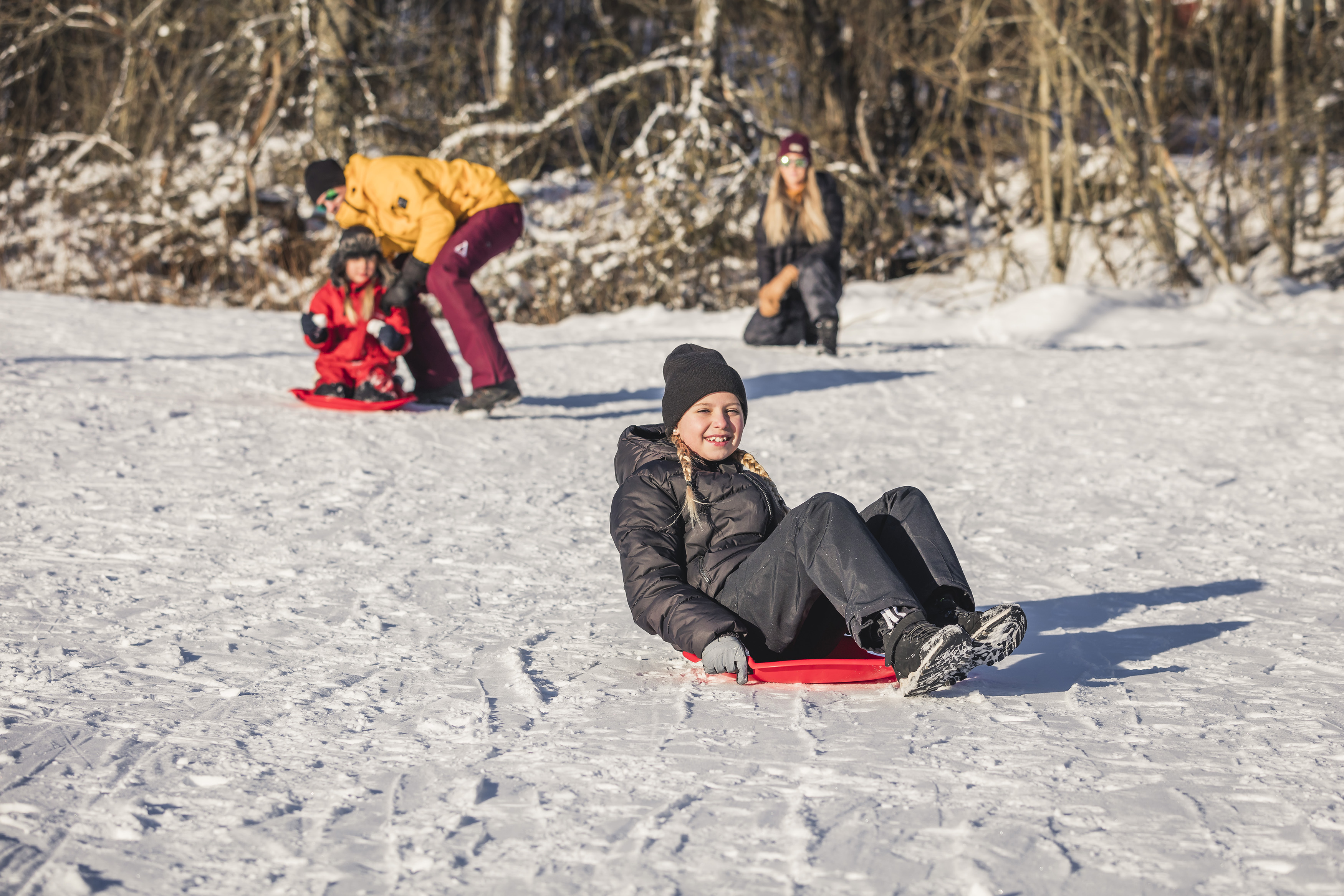 Girl riding on a red sleigh on the snow. Man, woman and a child in the background.