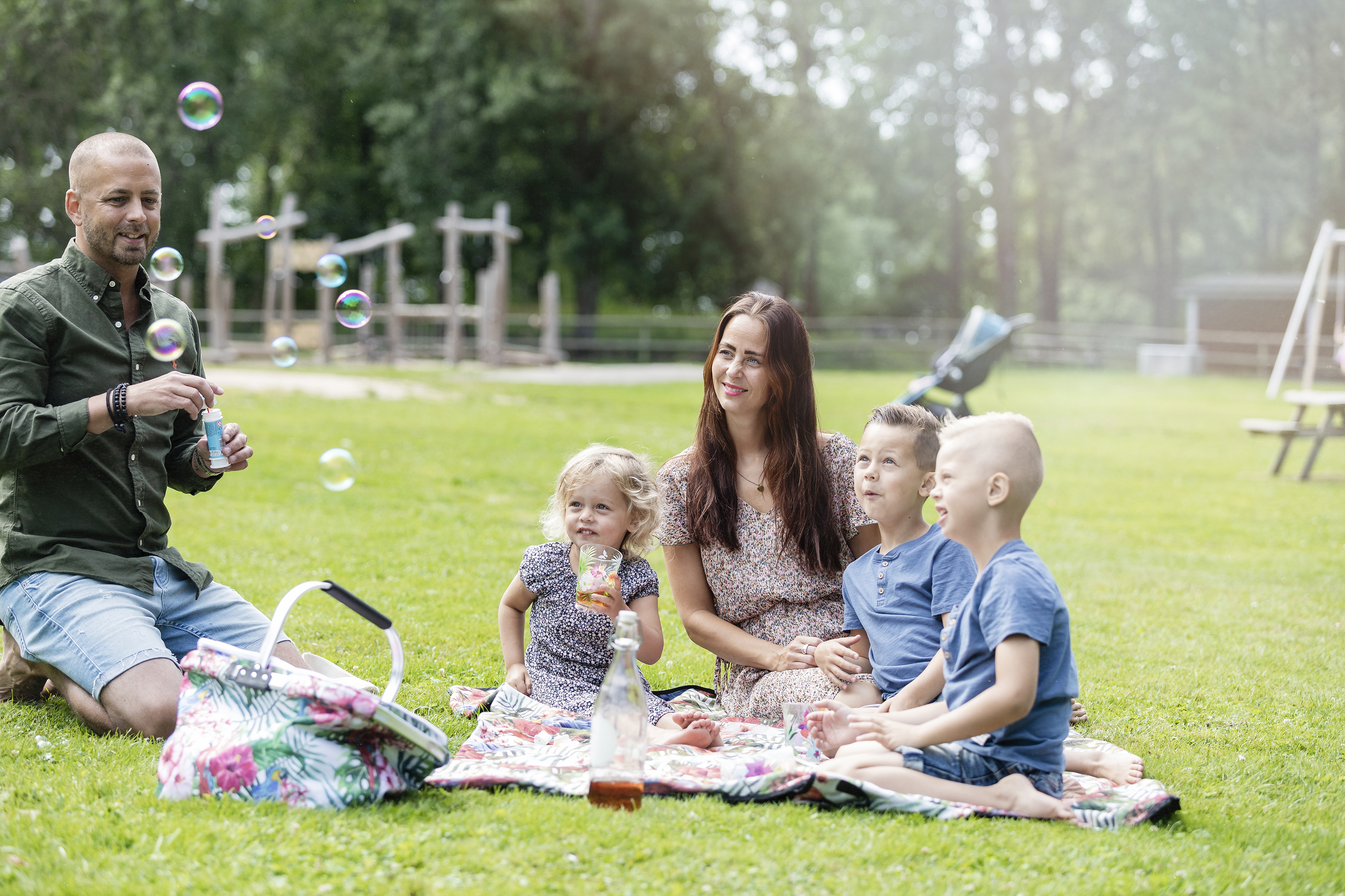 Pappa som blåser såpbubblor, tre barn och mamman sitter på en filt på gräset och ser på och fika.