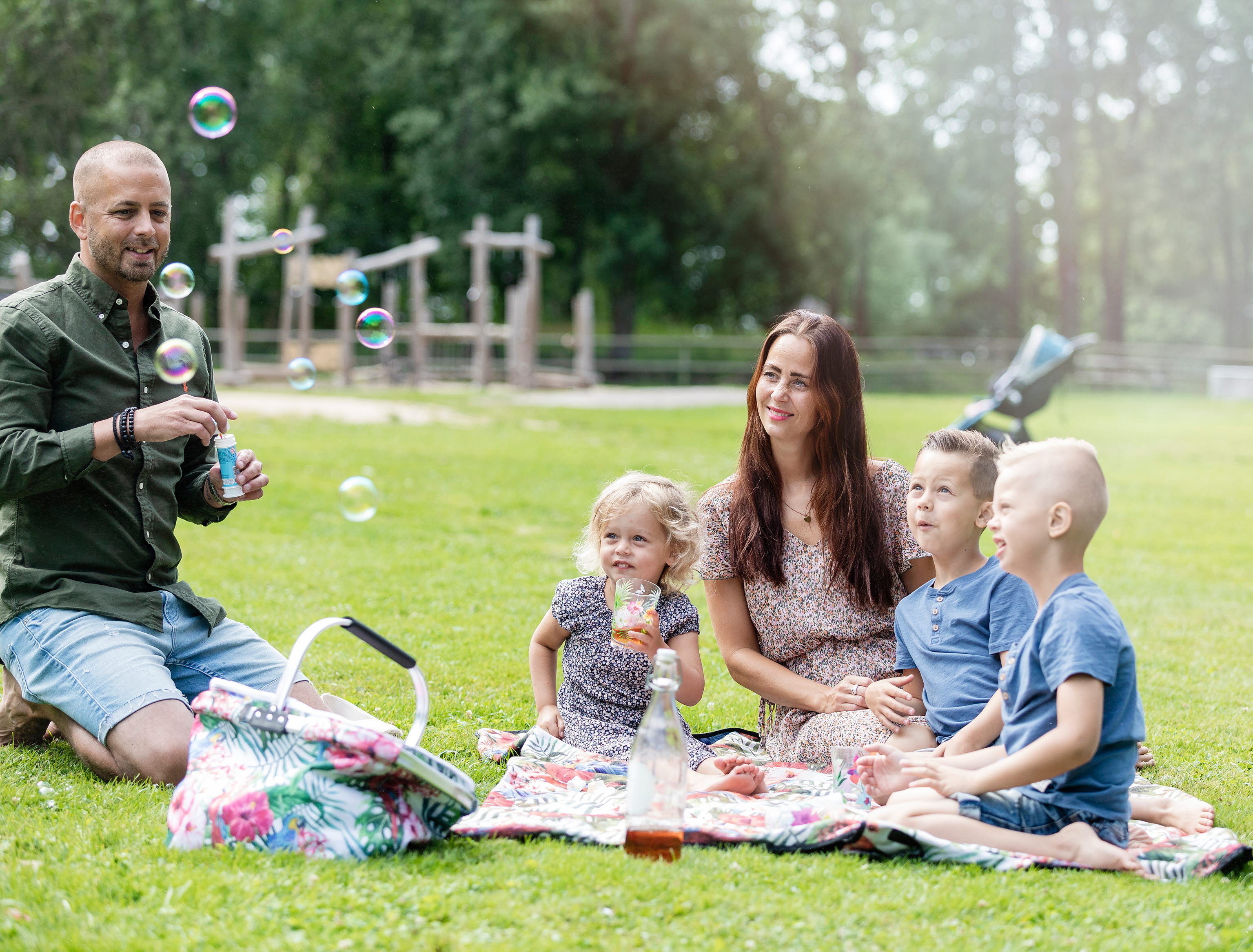 Pappa som blåser såpbubblor, tre barn och mamman sitter på en filt på gräset och ser på och fika.