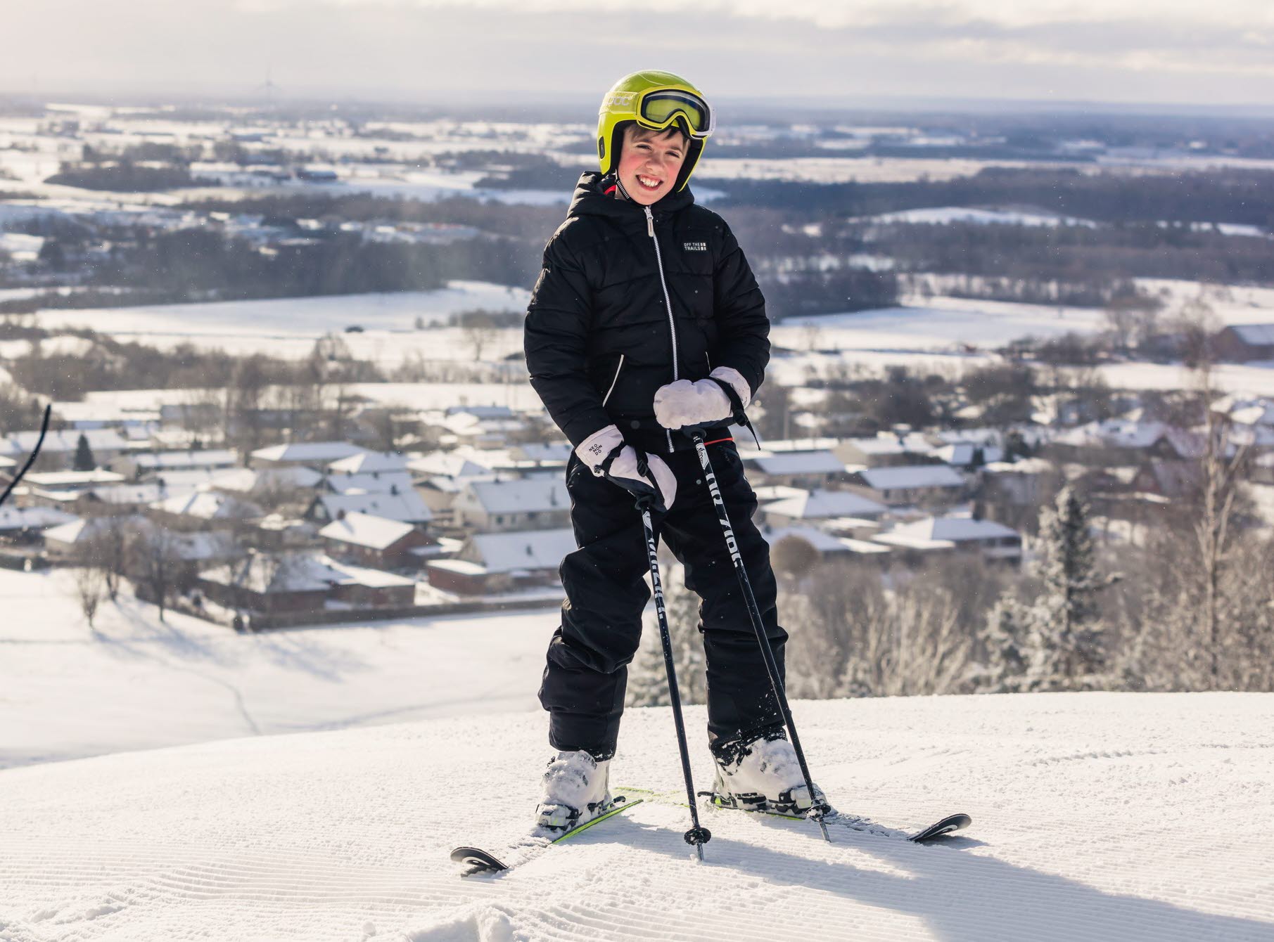 A child stands at the top of the ski slope with a view behind him. He has black ski clothes and a yellow helmet.
