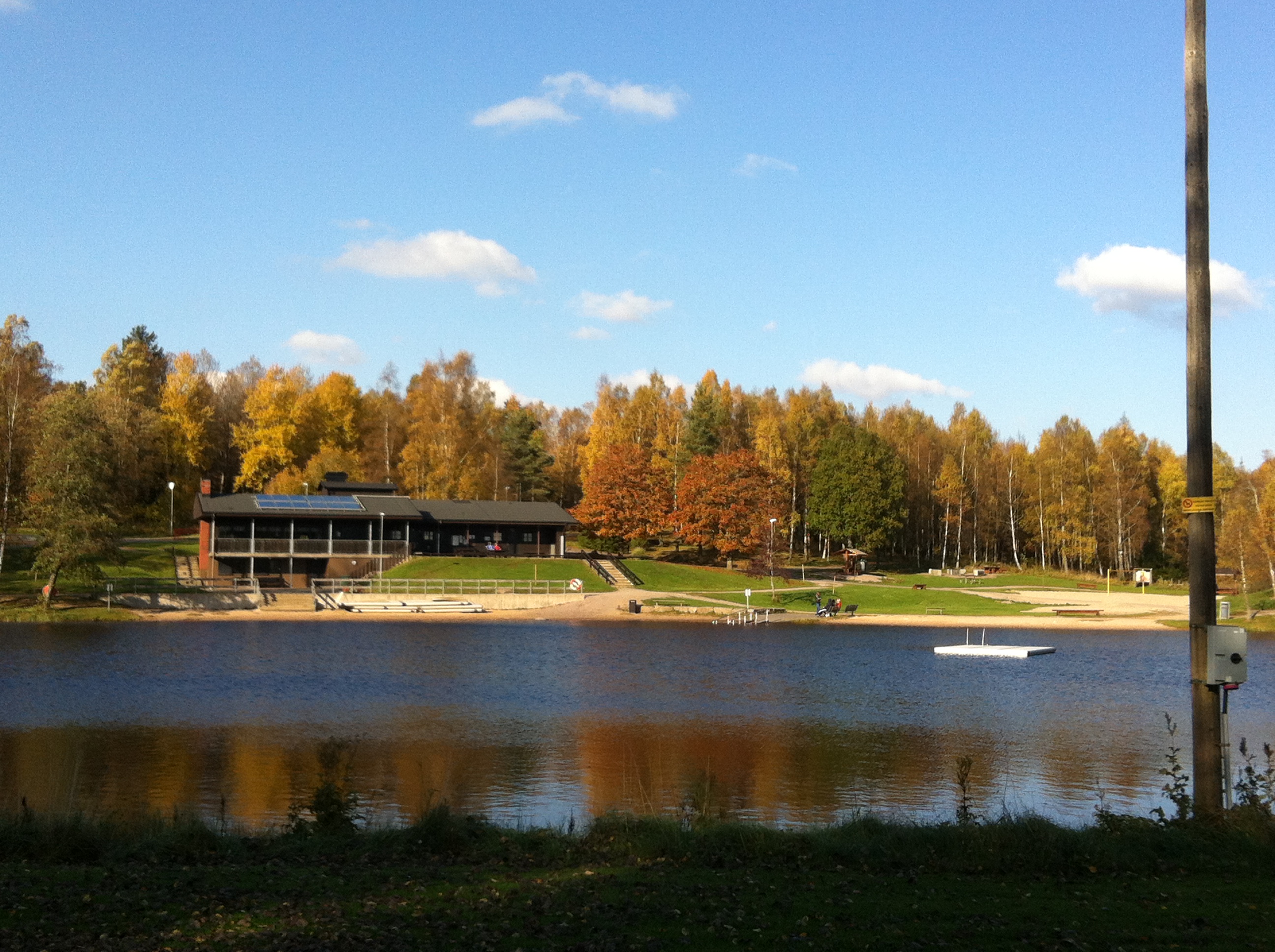 Byggnad i brunt trä och träd i höstfärger bredvid. Framför ligger en sjö.