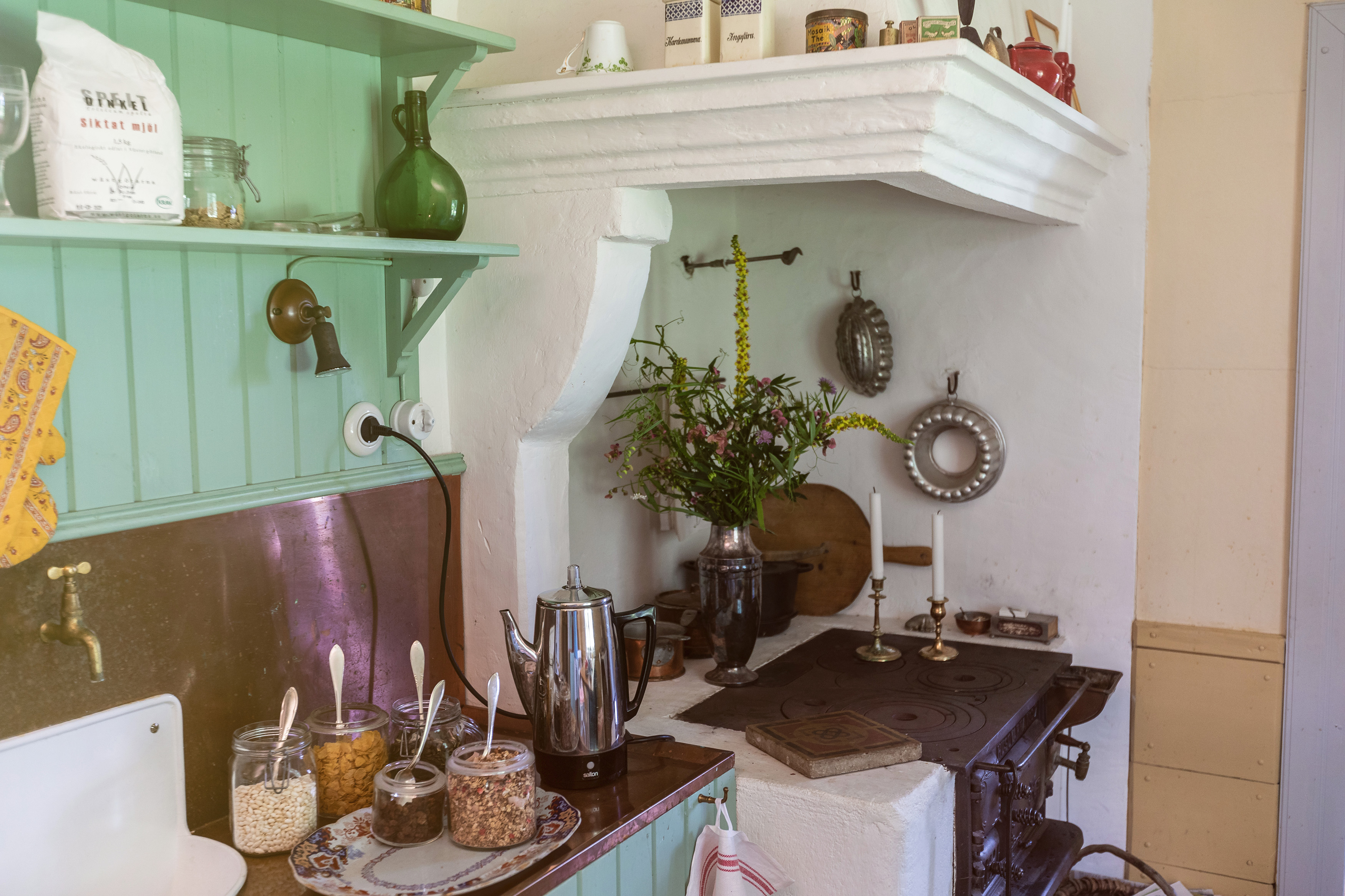 Ett gammaldags kök med vedspis och gröna träväggar. Frukostflingor står framdukat.