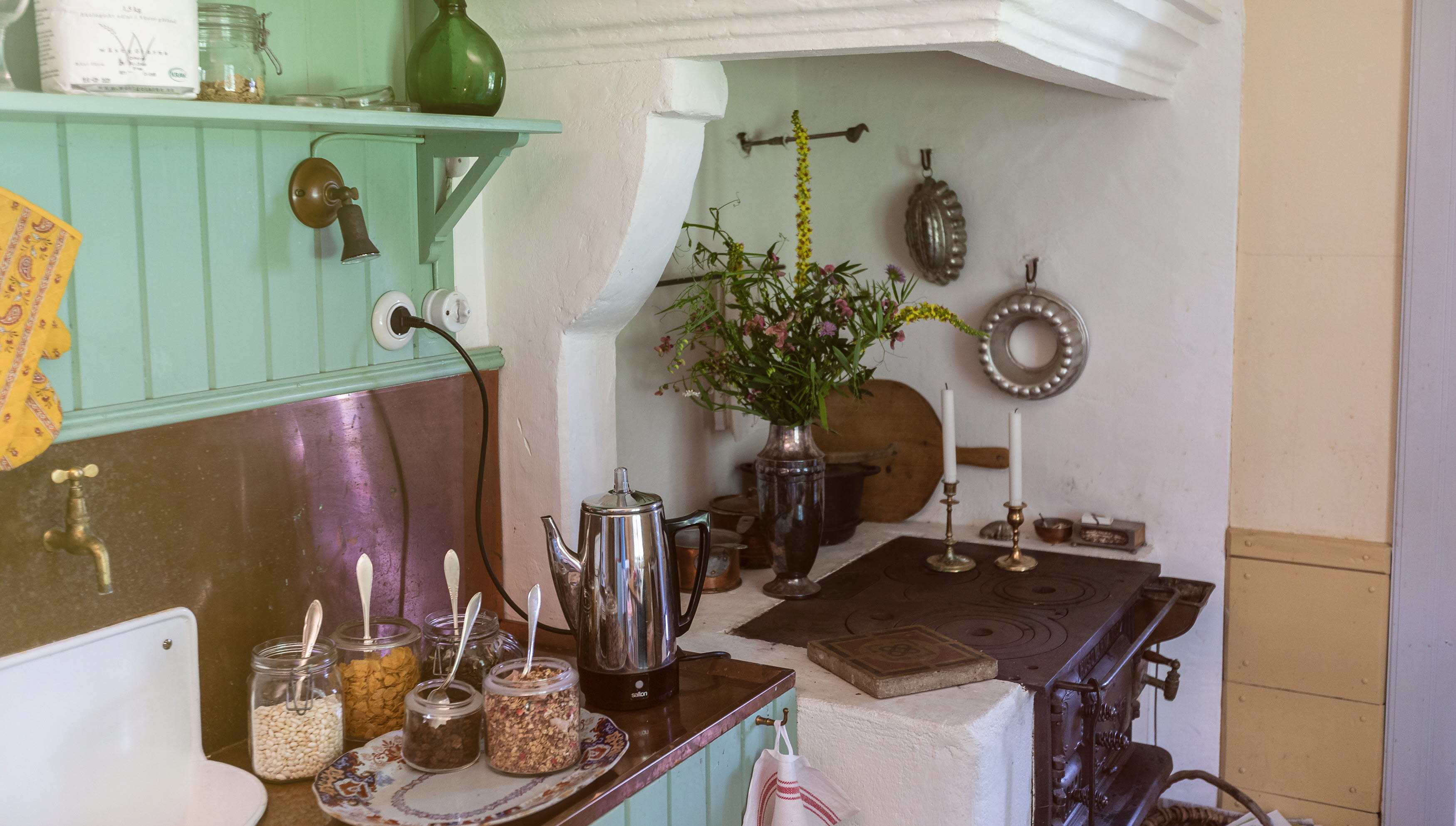 Ett gammaldags kök med vedspis och gröna träväggar. Frukostflingor står framdukat.