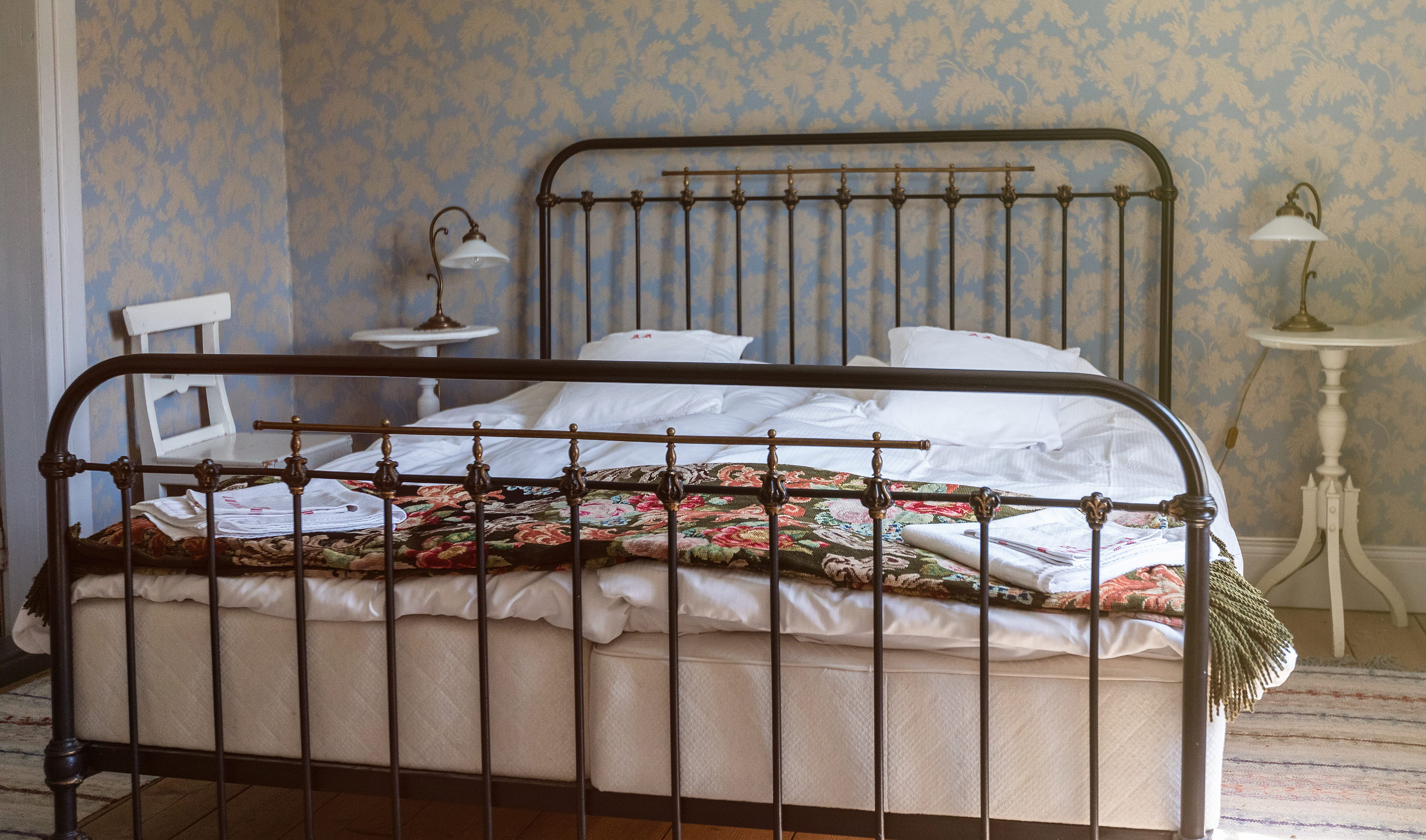 Dubbelsäng med järnram, blommiga taper i bakgrunden.