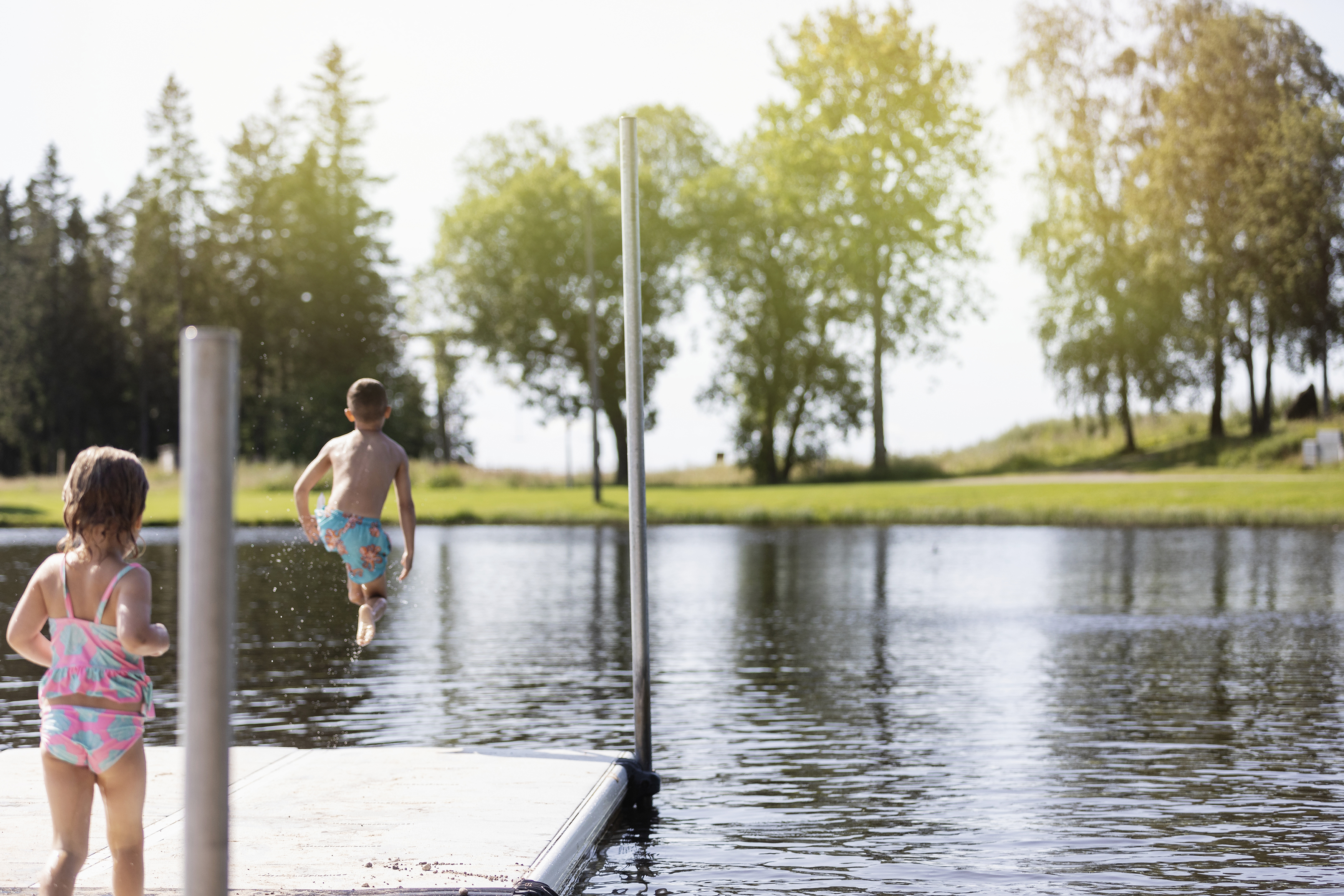 Två barn på en brygga med ryggen mot kameran. En av dem hoppar i vattnet.