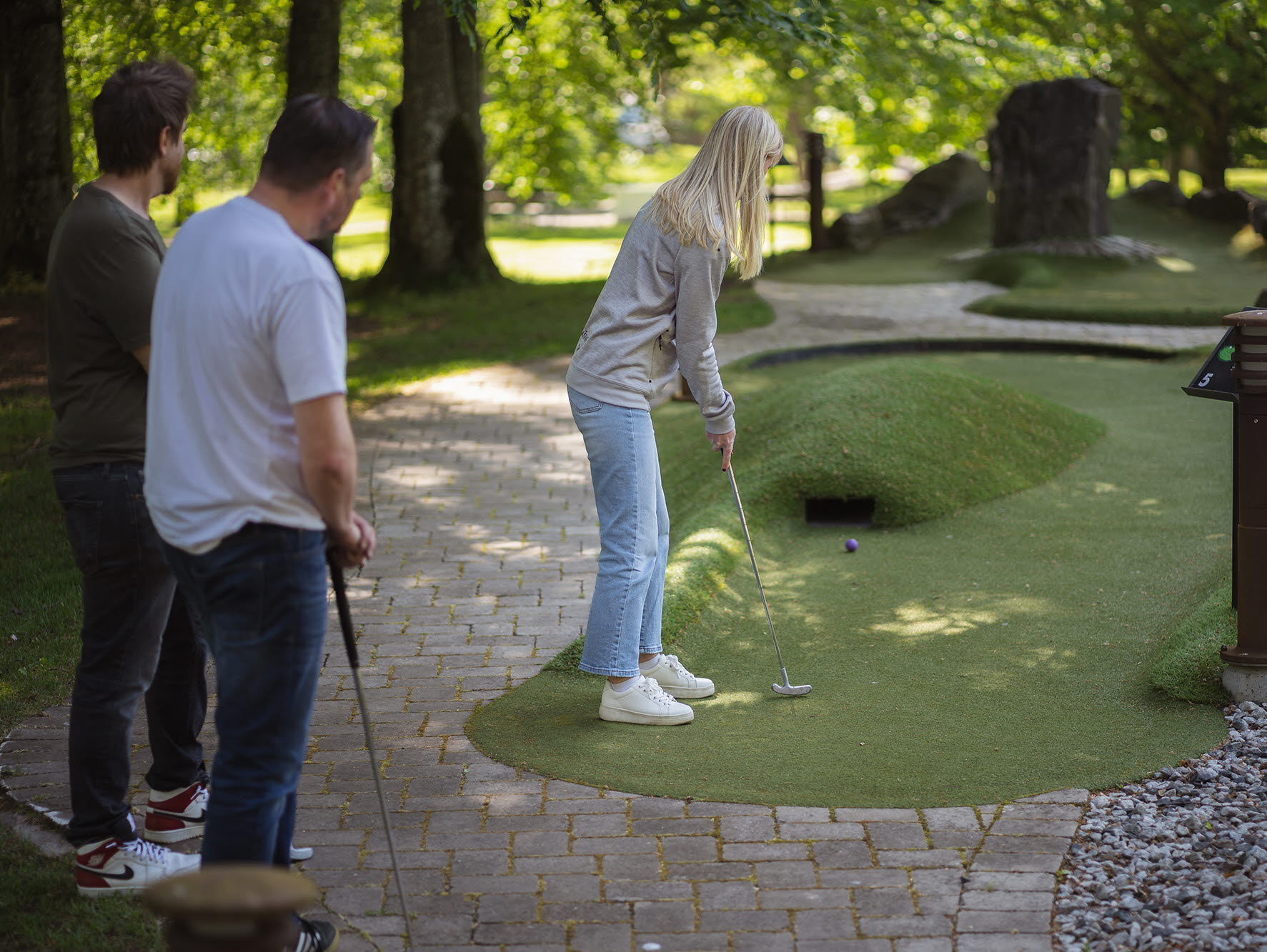 en person slår ett slag på minigolfbanan. I bakgrunden står två andra spelare och tittar på.