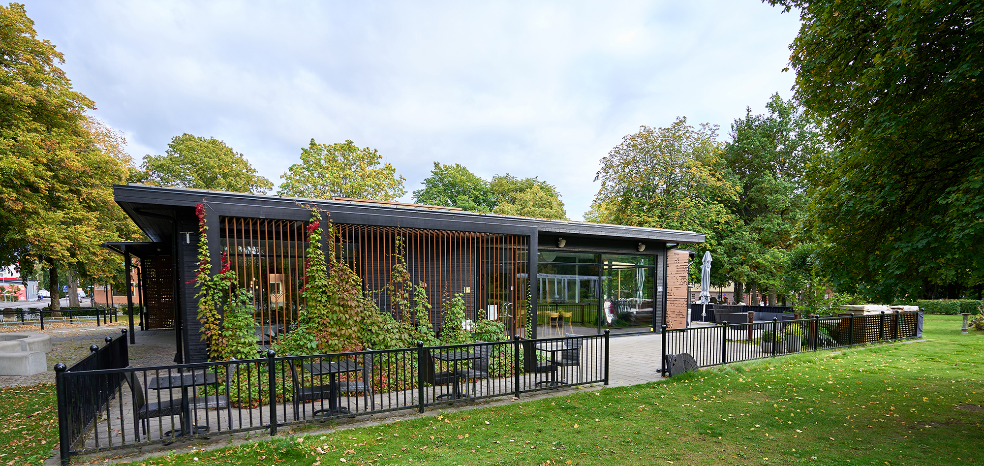 En byggnad med platt tak i svart och rostfärgat. 
Svart staket i metall runt om. En klätterväxt växer i på en pergola.