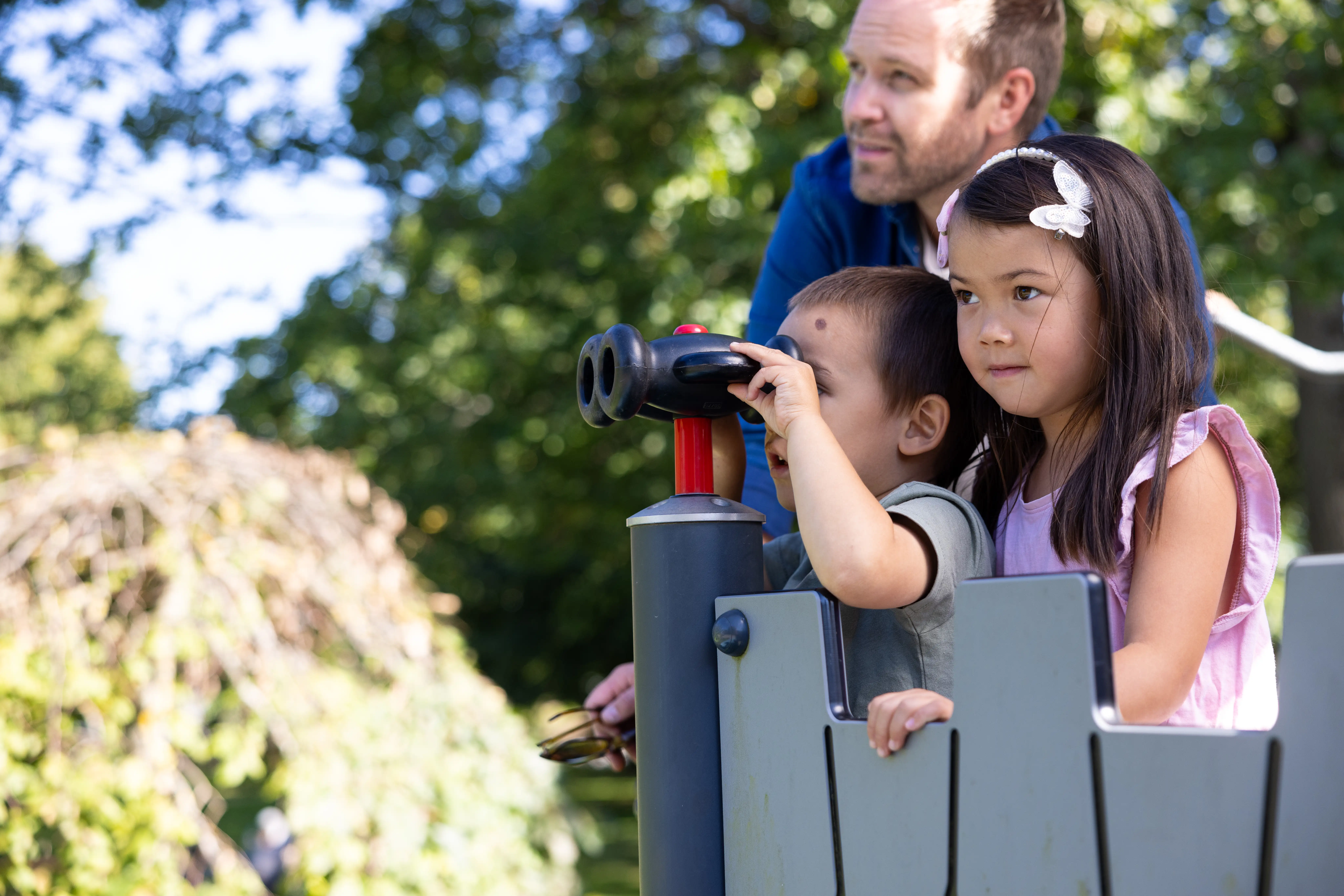 Två barn och en vuxen står i ett trätorn på en lekplats. Det ena barnet kikar med en kikare,