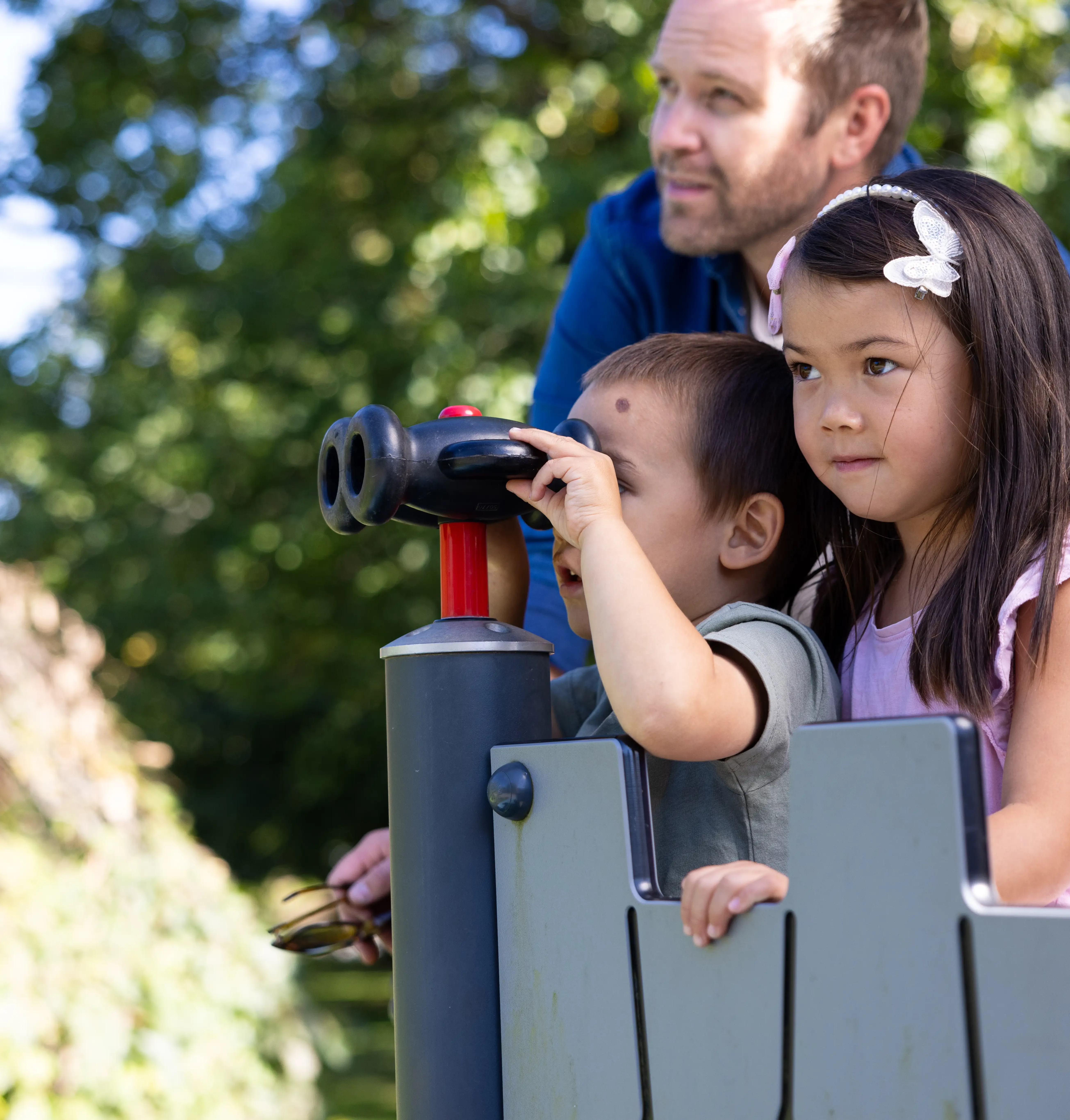 Två barn och en vuxen står i ett trätorn på en lekplats. Det ena barnet kikar med en kikare,