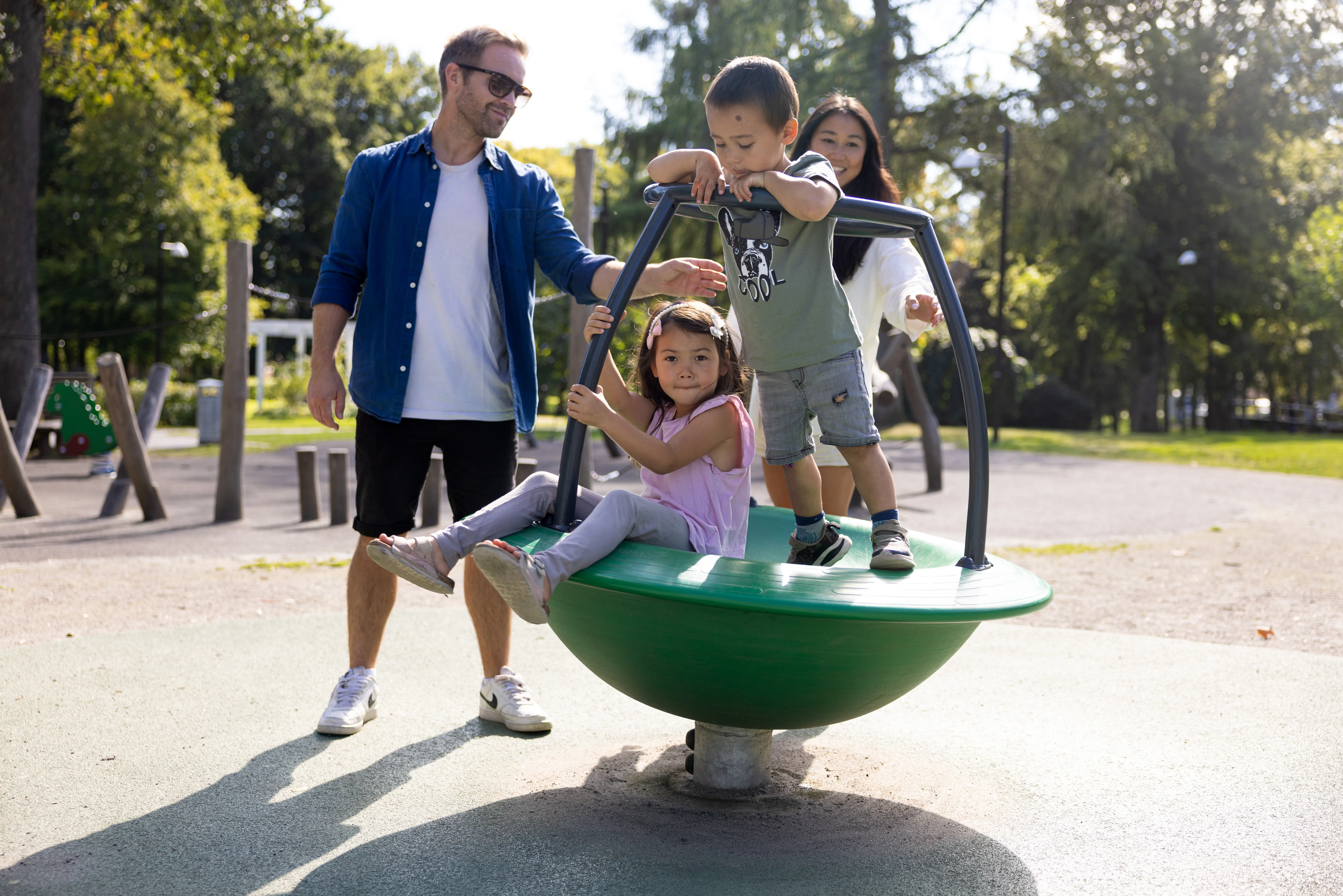två barn sitter och står i rund lekattraktion på en lekplats. Bredvid står två vuxna.