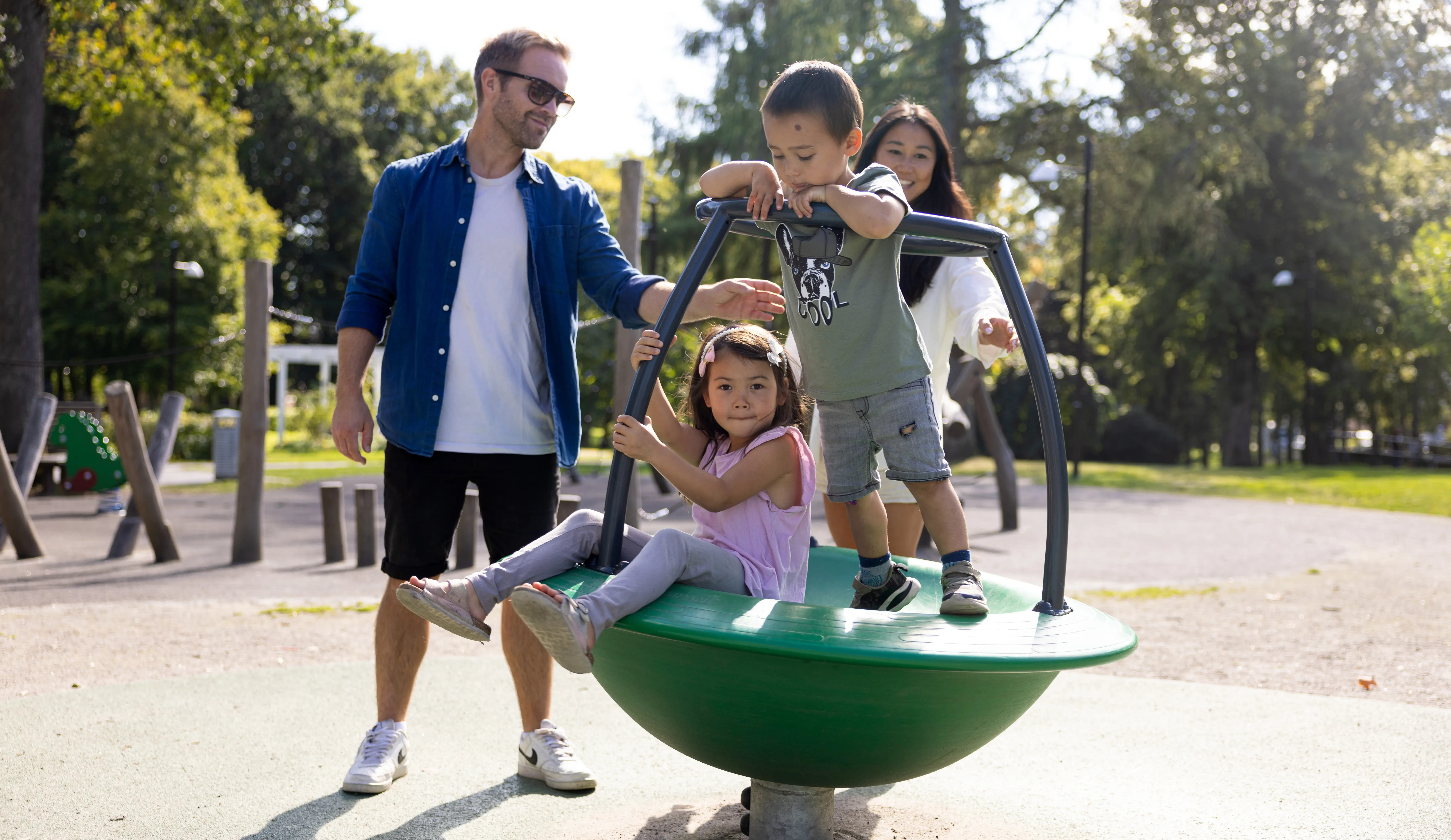 två barn sitter och står i rund lekattraktion på en lekplats. Bredvid står två vuxna.
