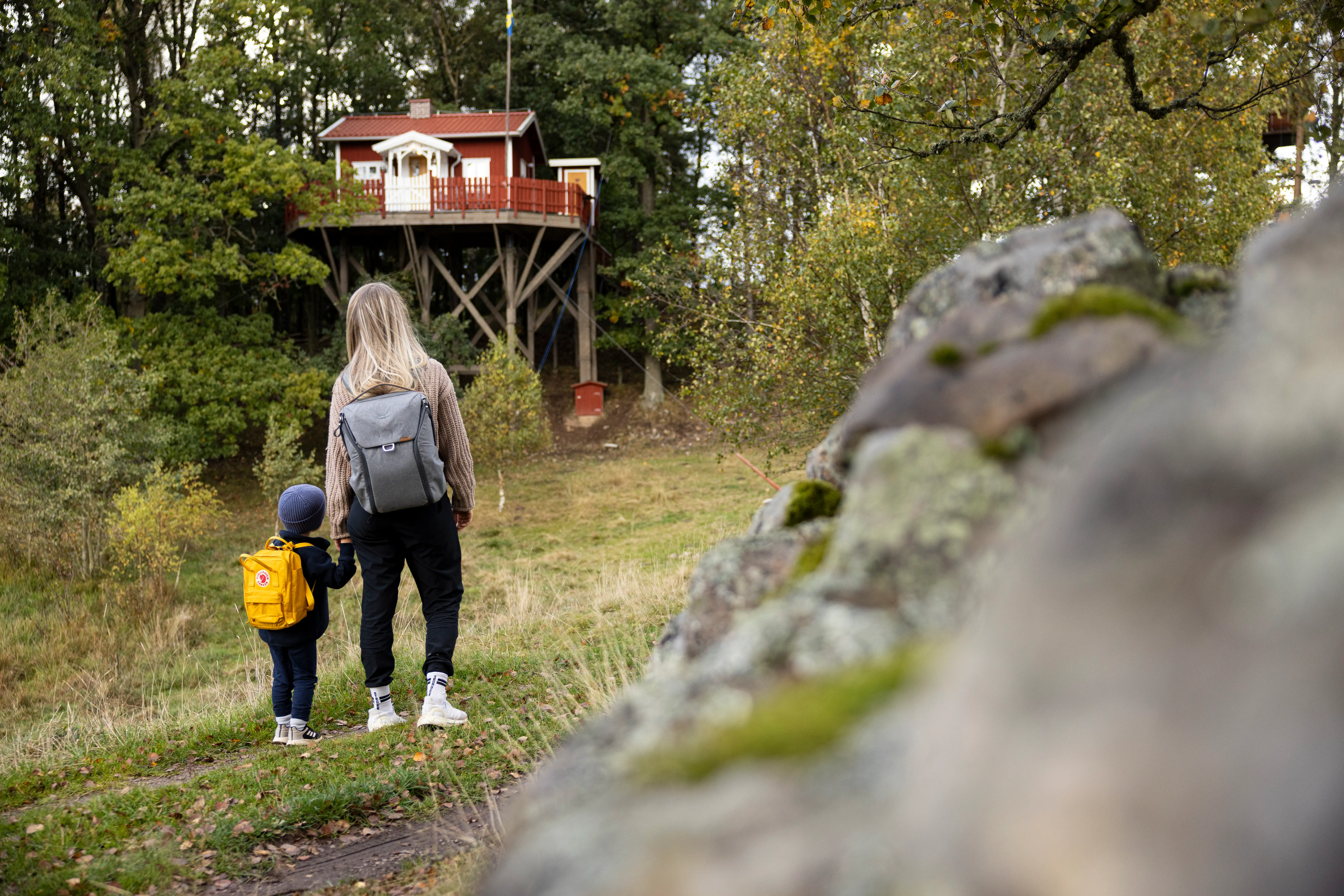 En kvinna och ett barn går nedanför ett trädhus. En röd stuga uppe i träden.