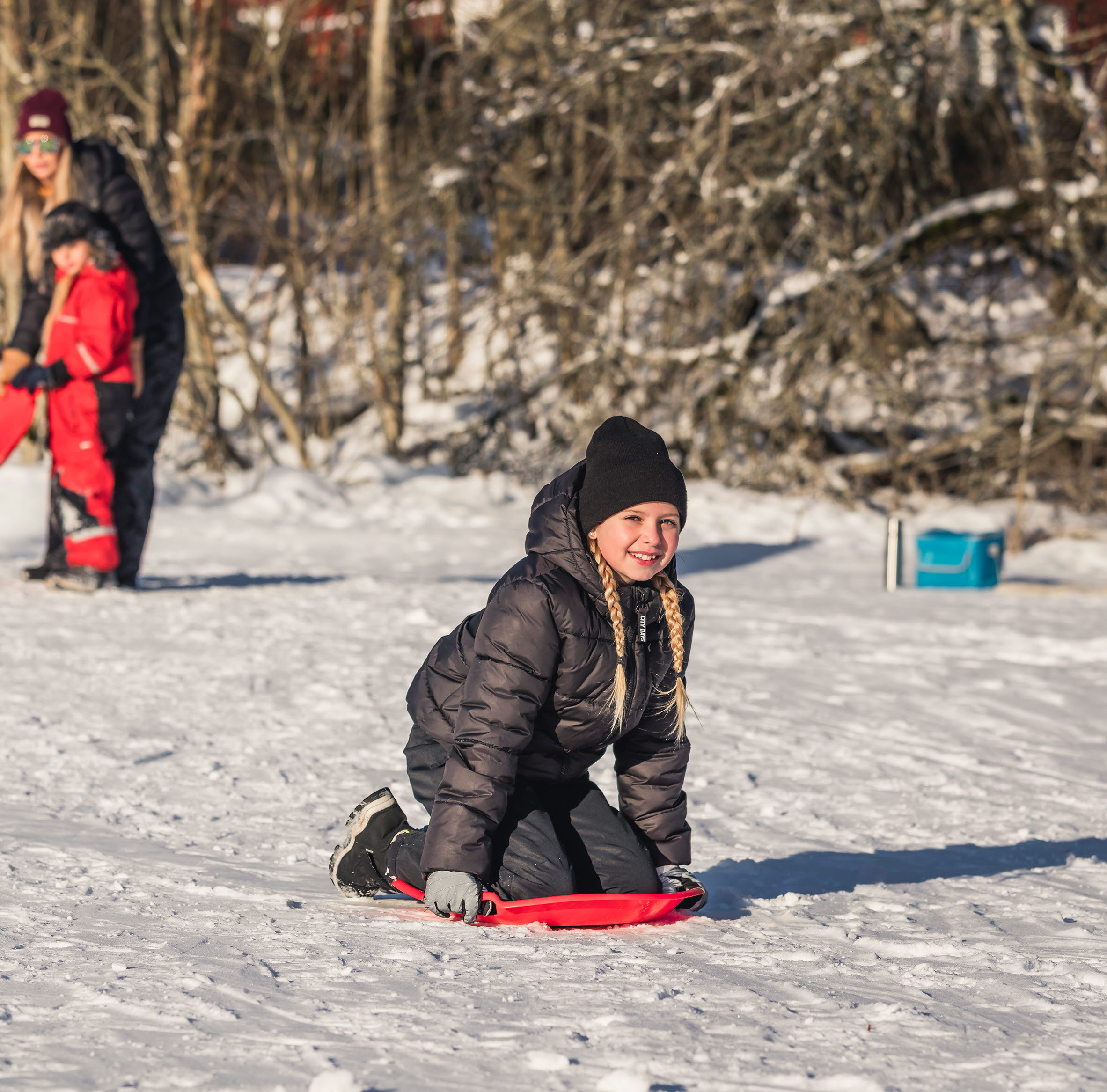 Flicka som sitter på knä på en röd pulka. Mamma och barn står i bakgrunden och tittar på.