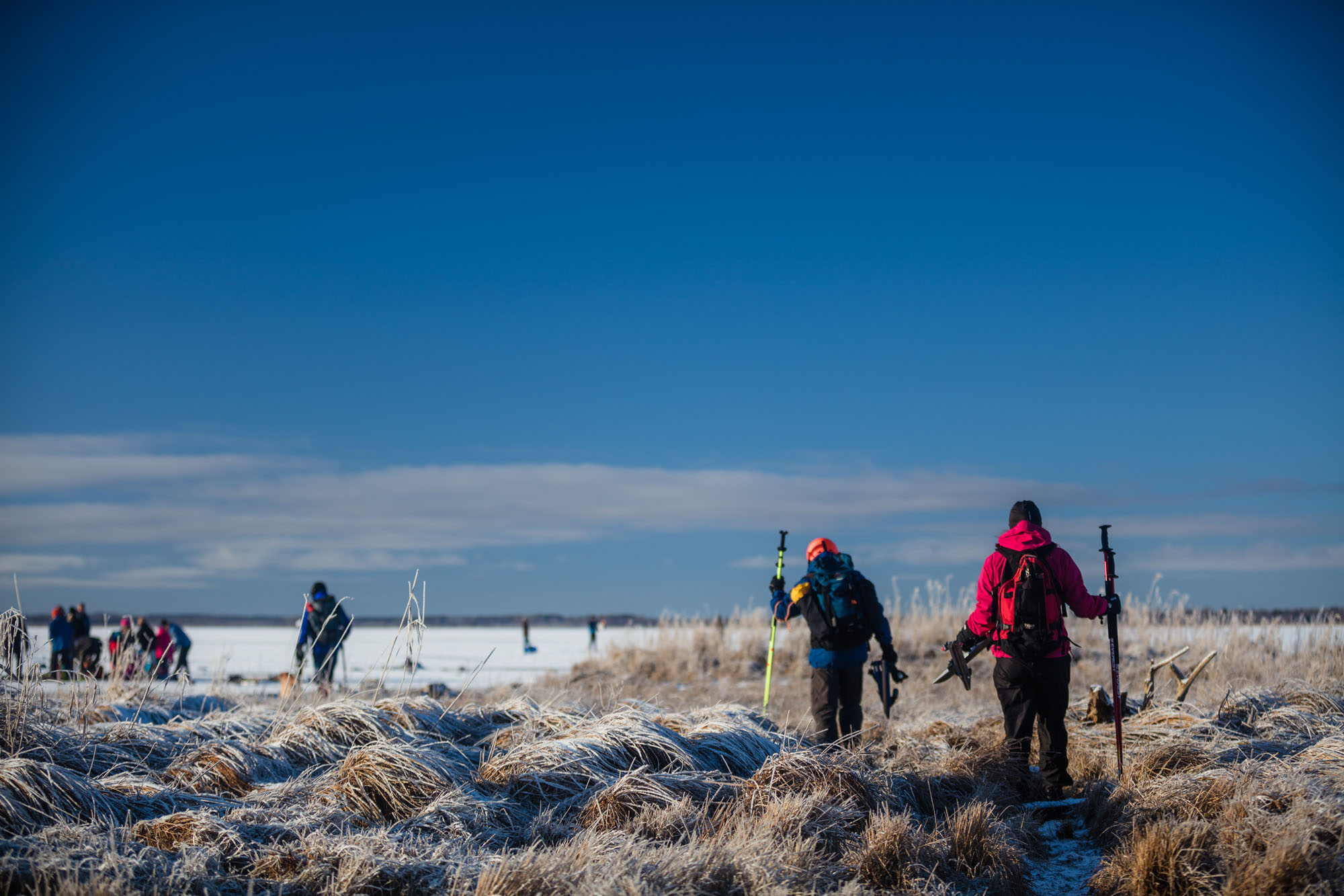 Två personer på väg ut till isen för att åka långfärdsskridskor. Blå himmel.
