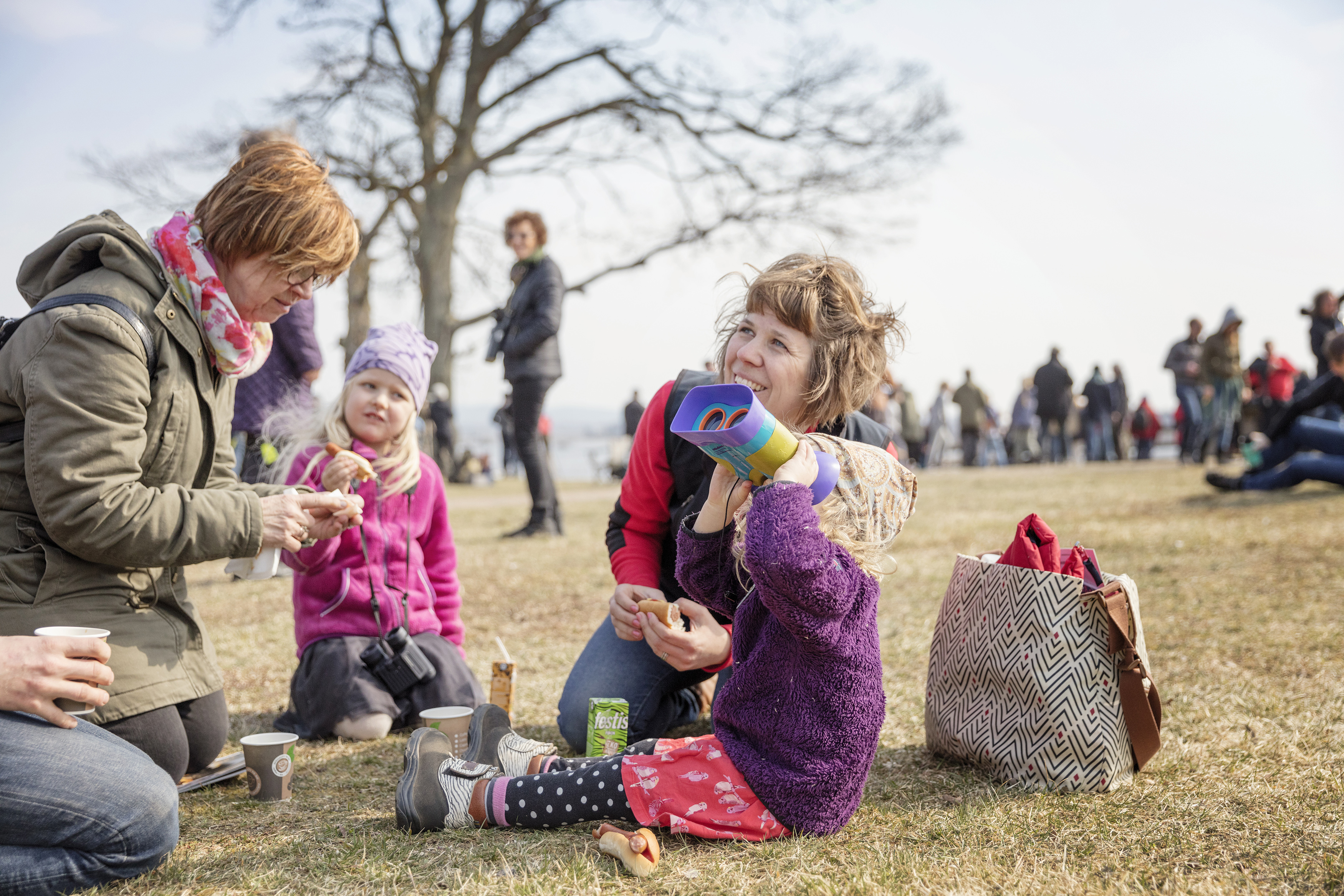 Barn och vuxna som sitter på marken och fikar. Ett av barnen kikar mot himlen med kikare.