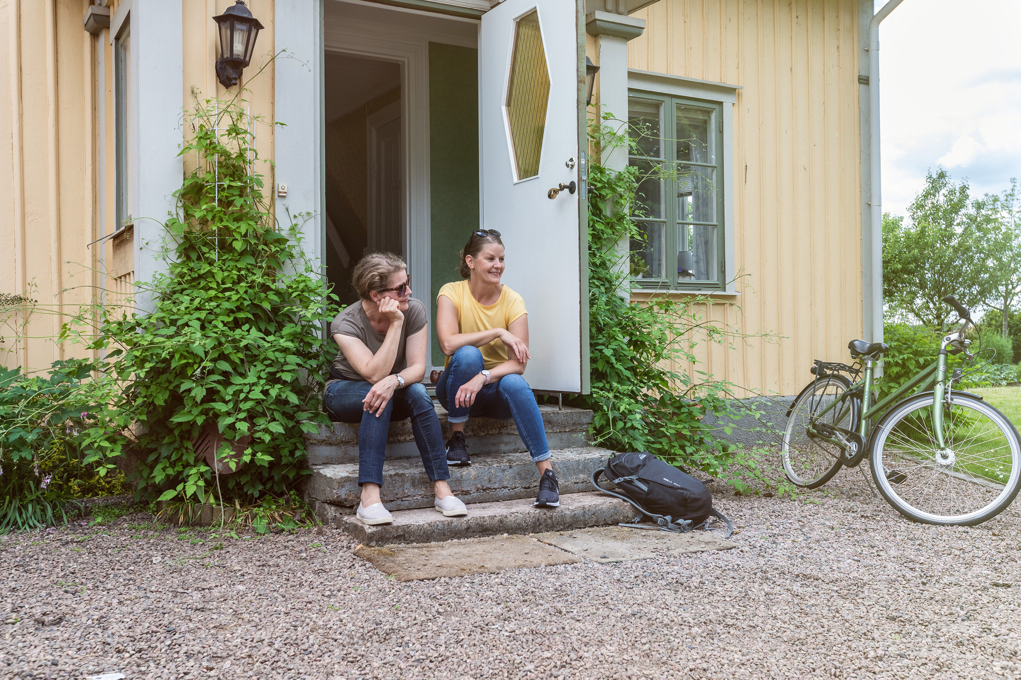 Två kvinnor sitter på en trappa till ett gult hus. Cykel står bredvid.