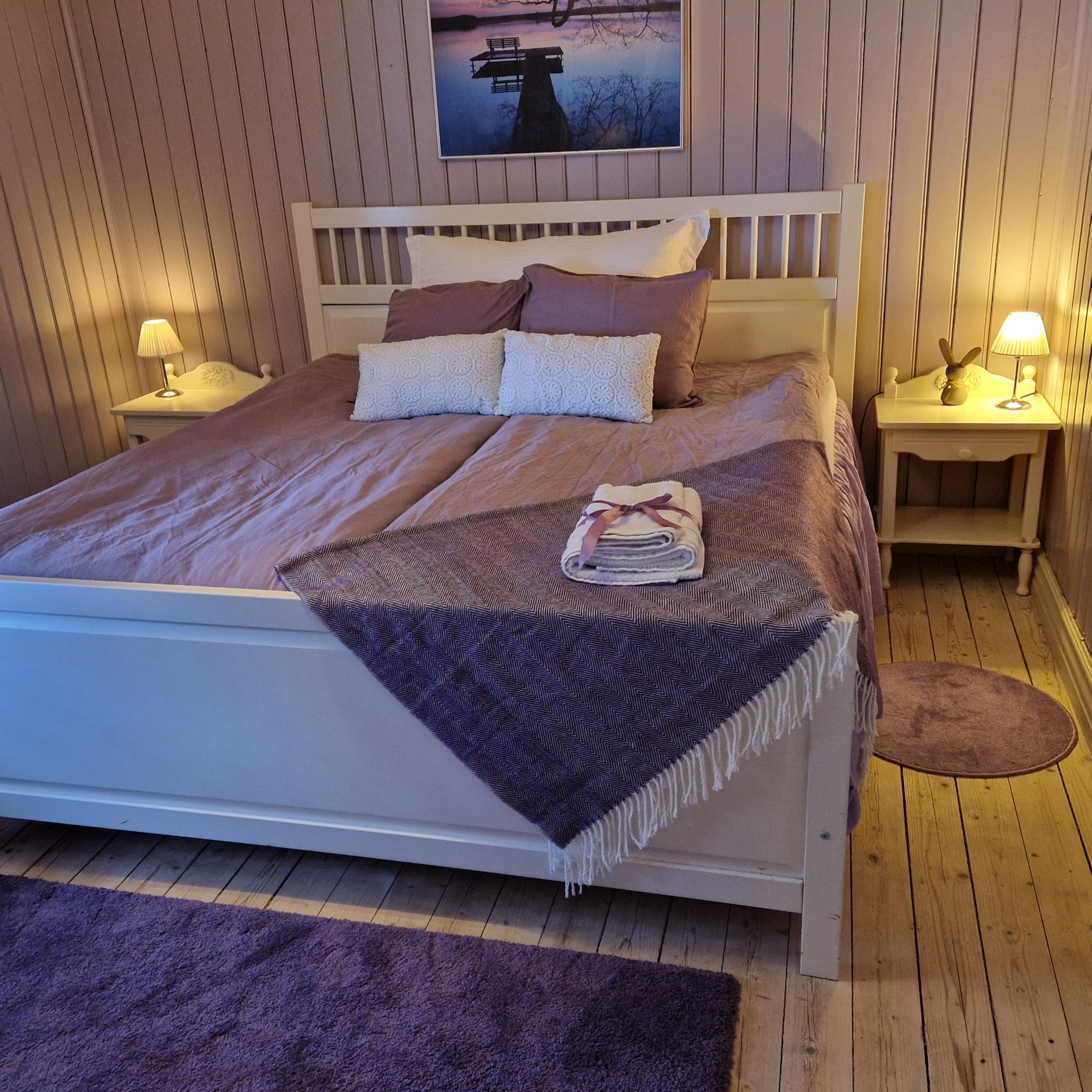 Vit säng med lila påslakanm kuddar och filt.