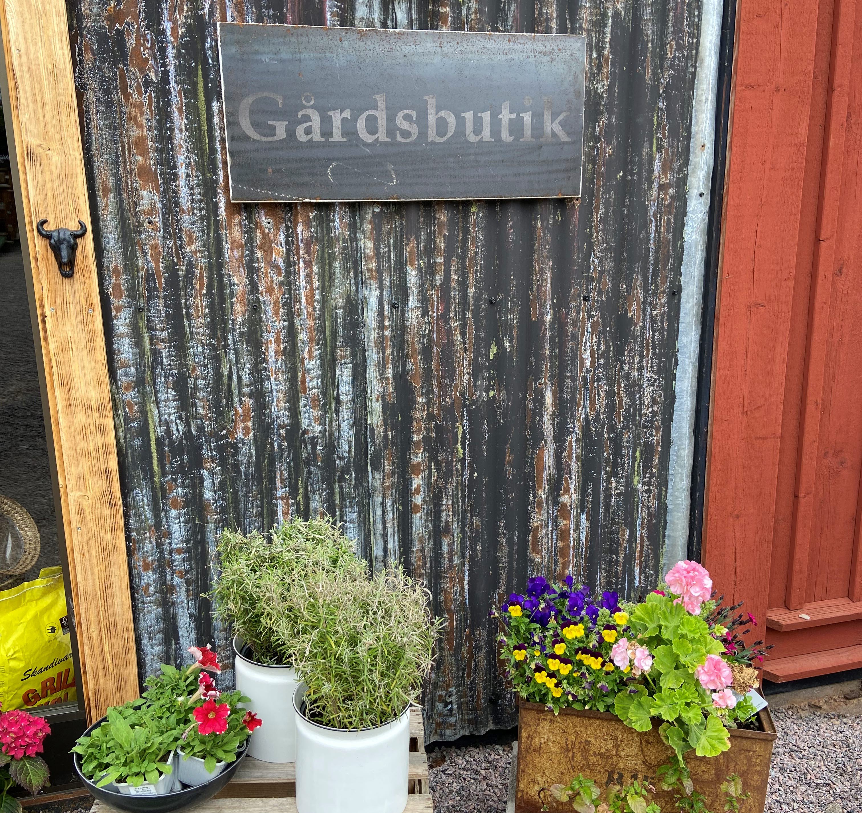 Svart plåtvägg med en skylt med texten Gårdsbutik. Blommor står nedanför skylten i krukor.