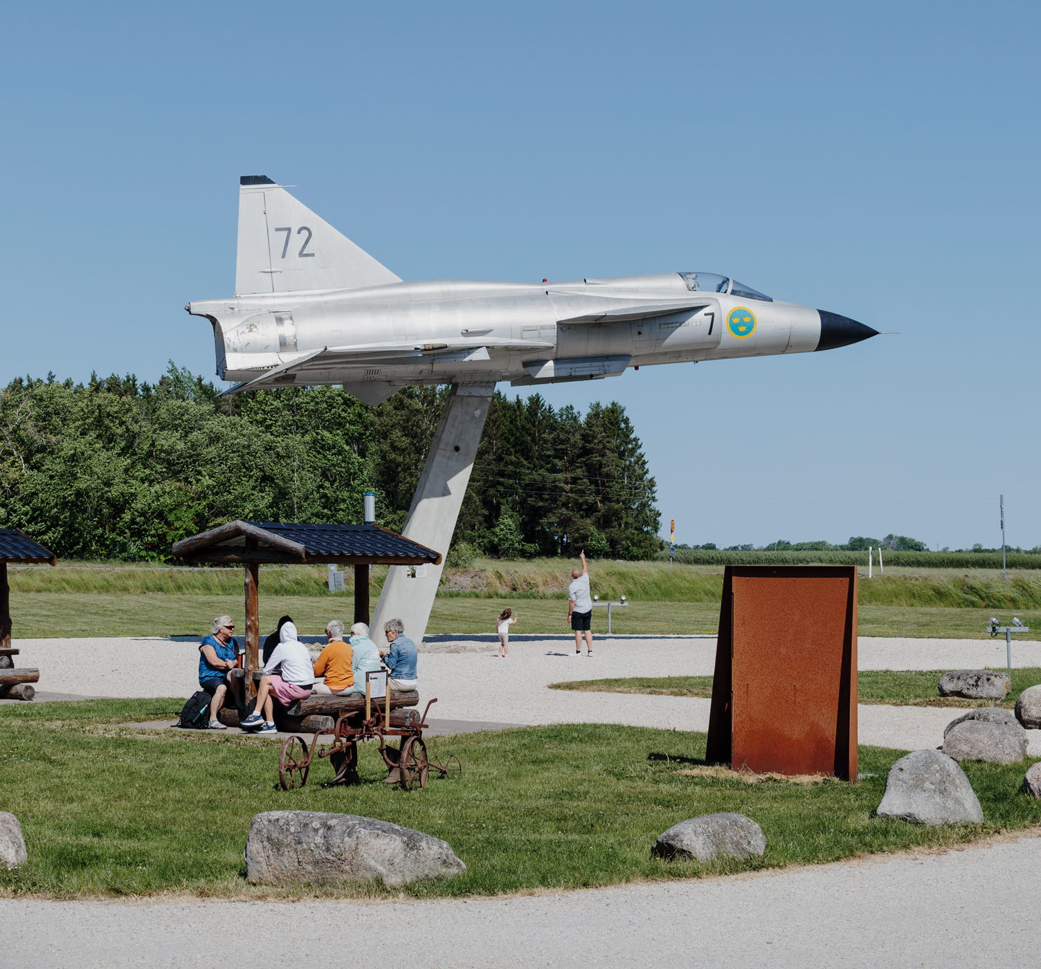 Rastplats Viggen i Grästorp med stort flygplan. Människor sitter och fikar vid ett bord. En förälder och ett barn tittar på flygplanet och pekar