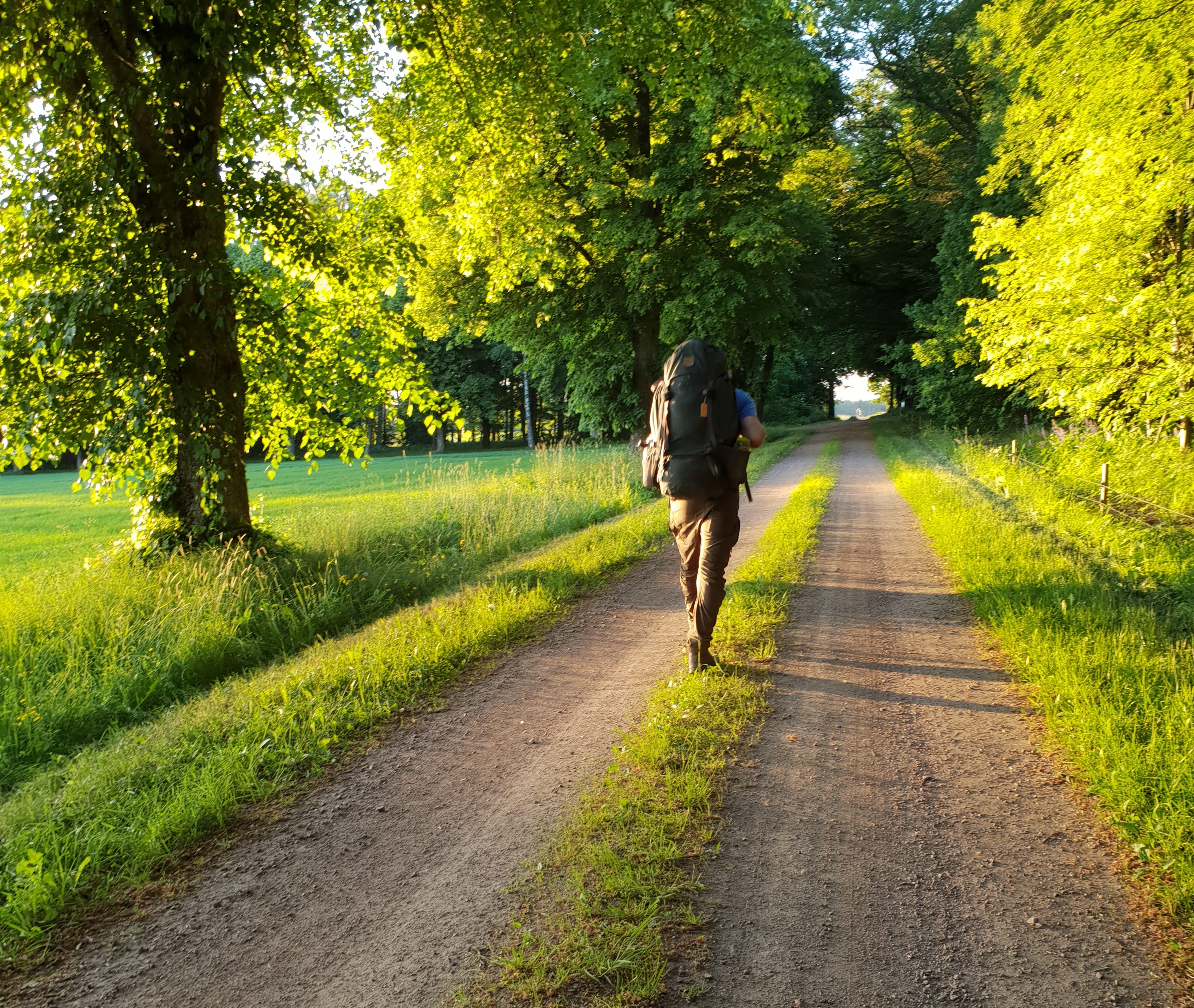 En kille med vandringsryggsäck går på grusväg i kvällsolen.