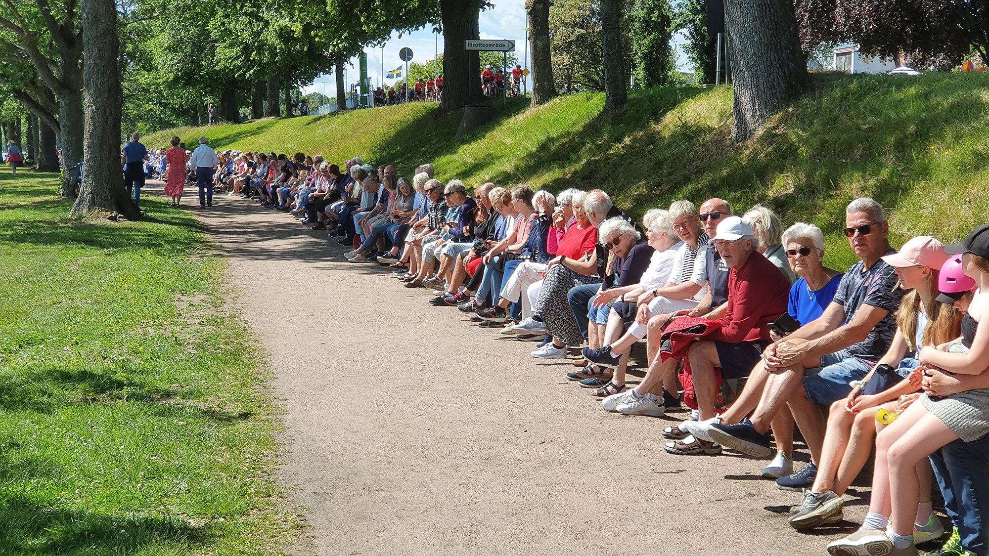 Människor sitter på Sveriges längsta bänk i Hjo, på Strandpromenaden en solig sommardag.