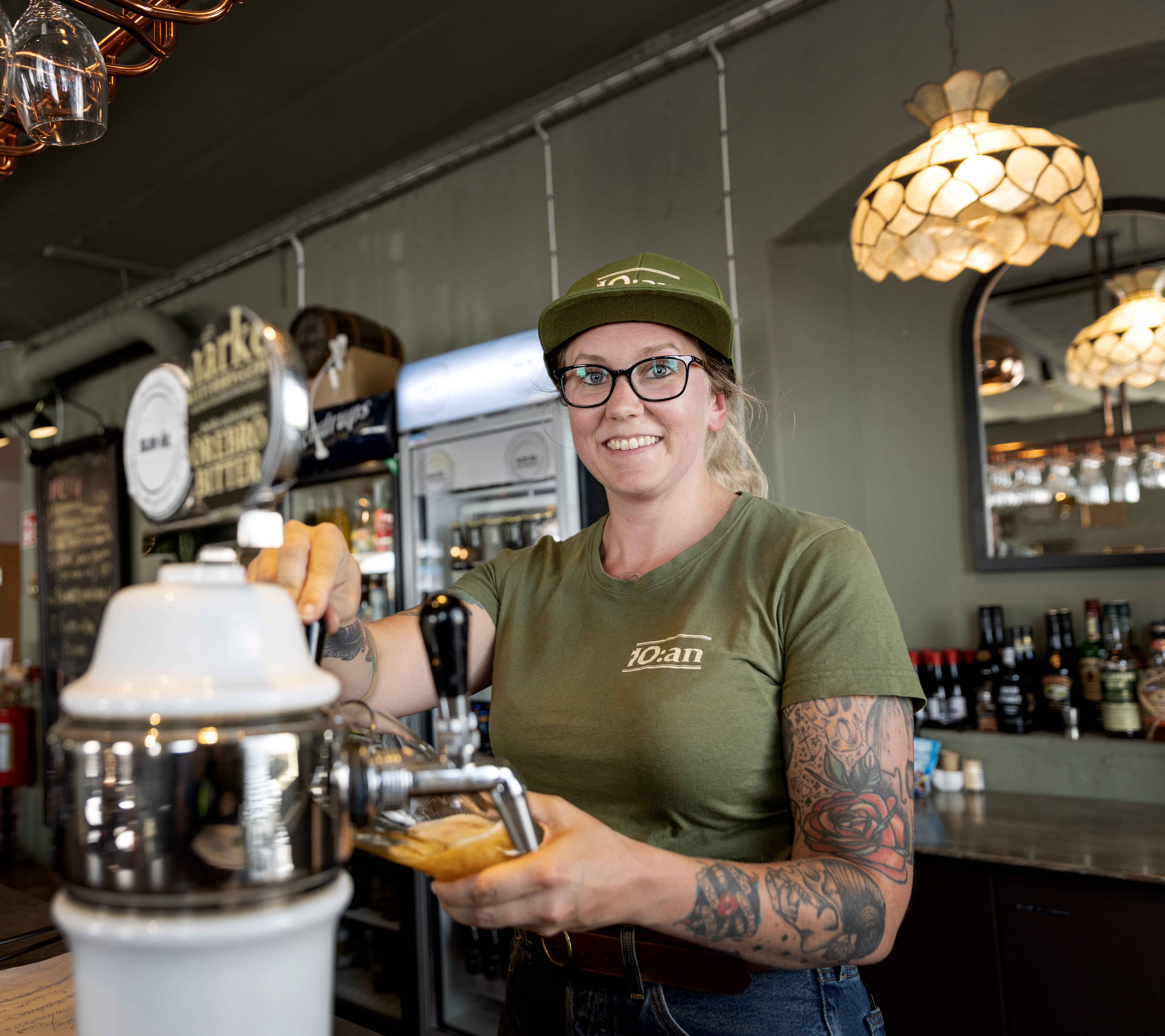 Tjej i grön tröja och keps står i en bar och häller upp en öl ur en tapp. glas hänger i taket. 