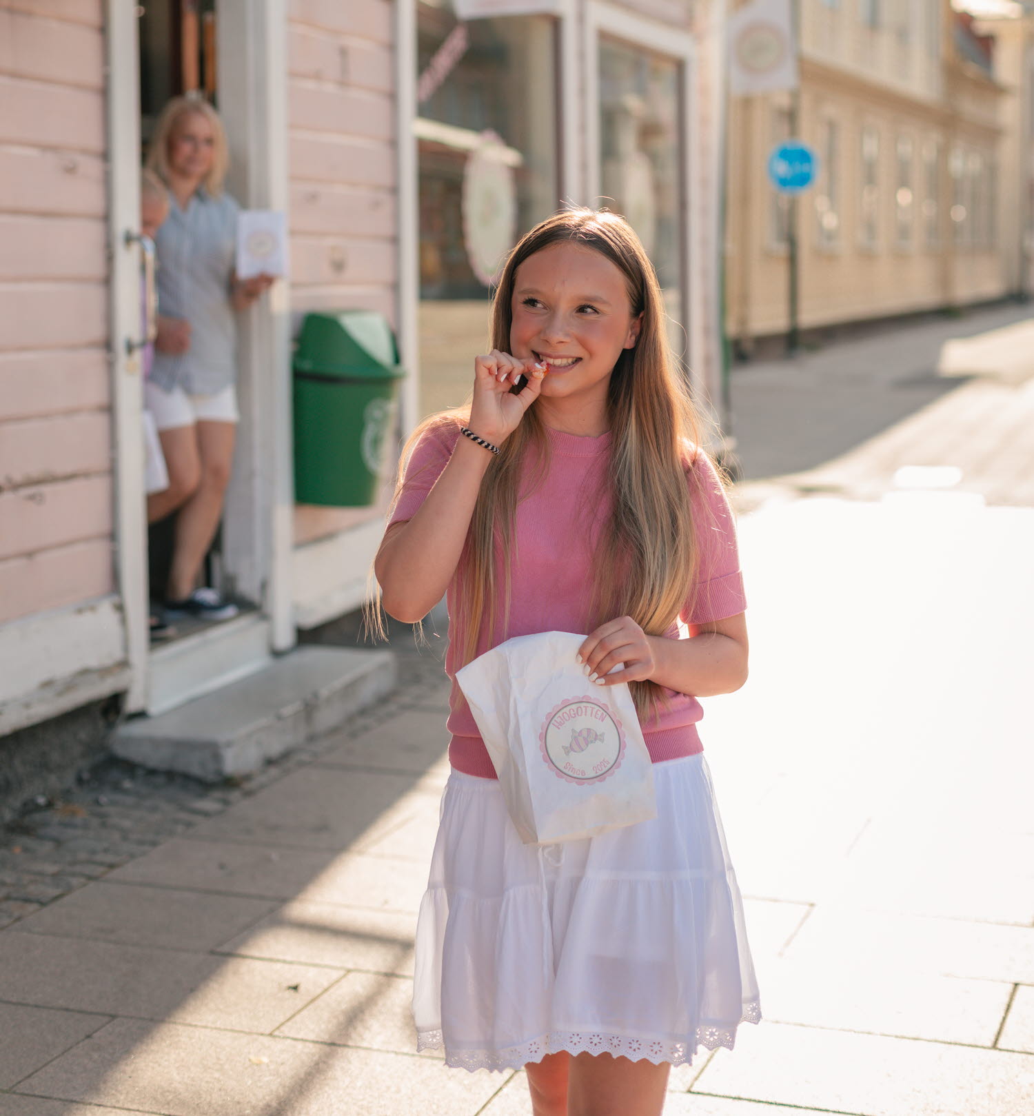 En tjej kommer ut från Hjogotten i bakgrunden, i förgrunden biter en annan tjej ned i en godisbit handlad på samma ställe