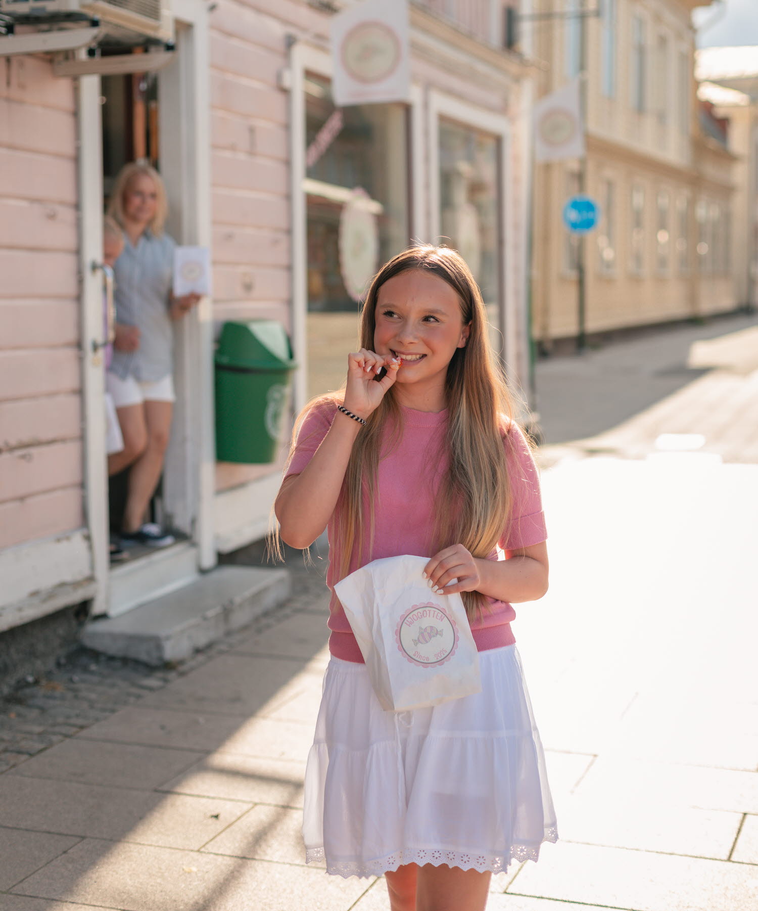 En tjej kommer ut från Hjogotten i bakgrunden, i förgrunden biter en annan tjej ned i en godisbit handlad på samma ställe