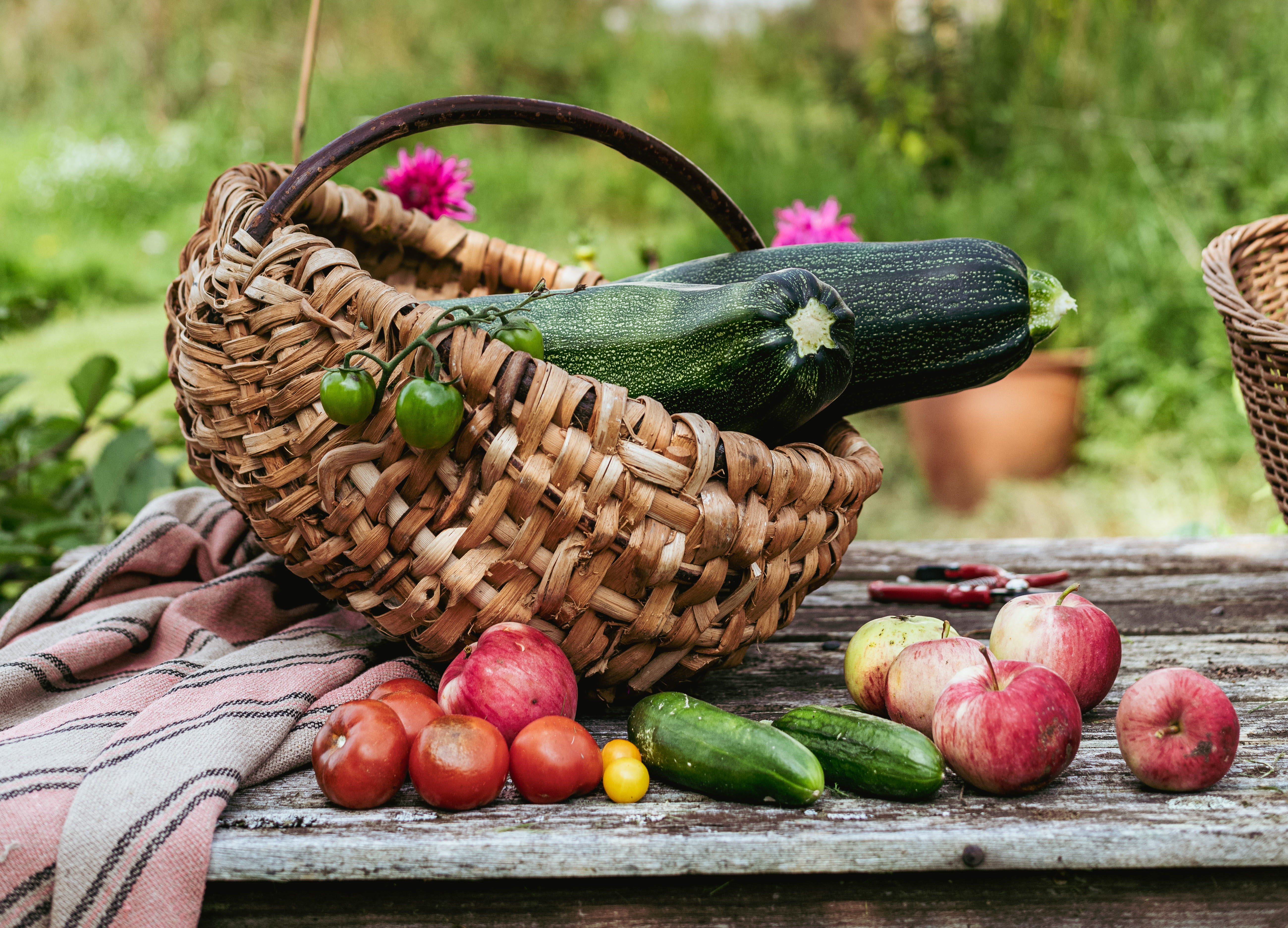 Squash i en korg, äpplen på ett äldre träbord bordet tillsammans med små gurkor och små röda tomater. Ett vitt stenhus i bakgrunden med en röd gammal dubbeldörr. 
