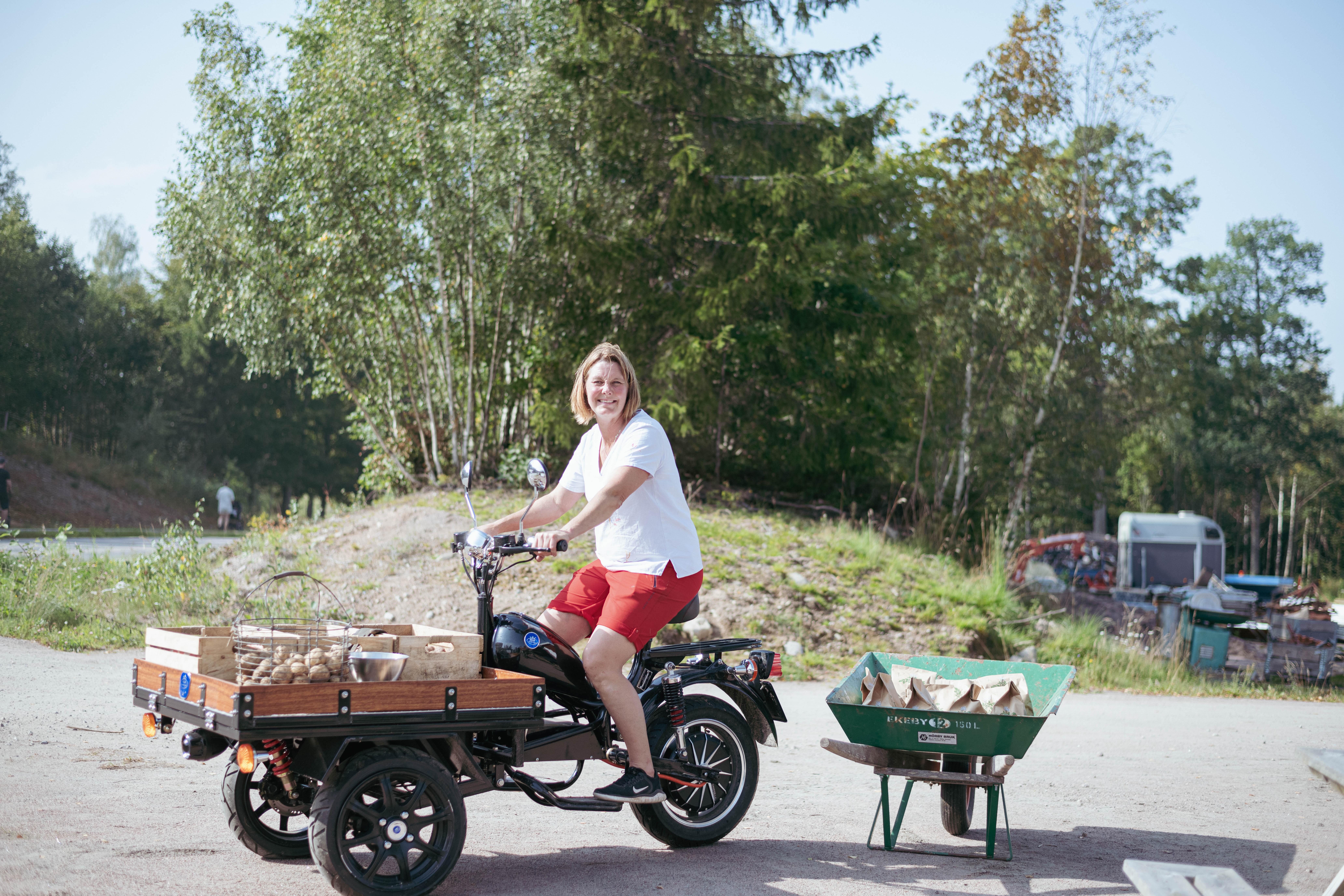 kvinna med röda shorts sitter på en flakmoppe  som är fylld med potatis. Bakom henne står en grön skottkärra med potatis. 