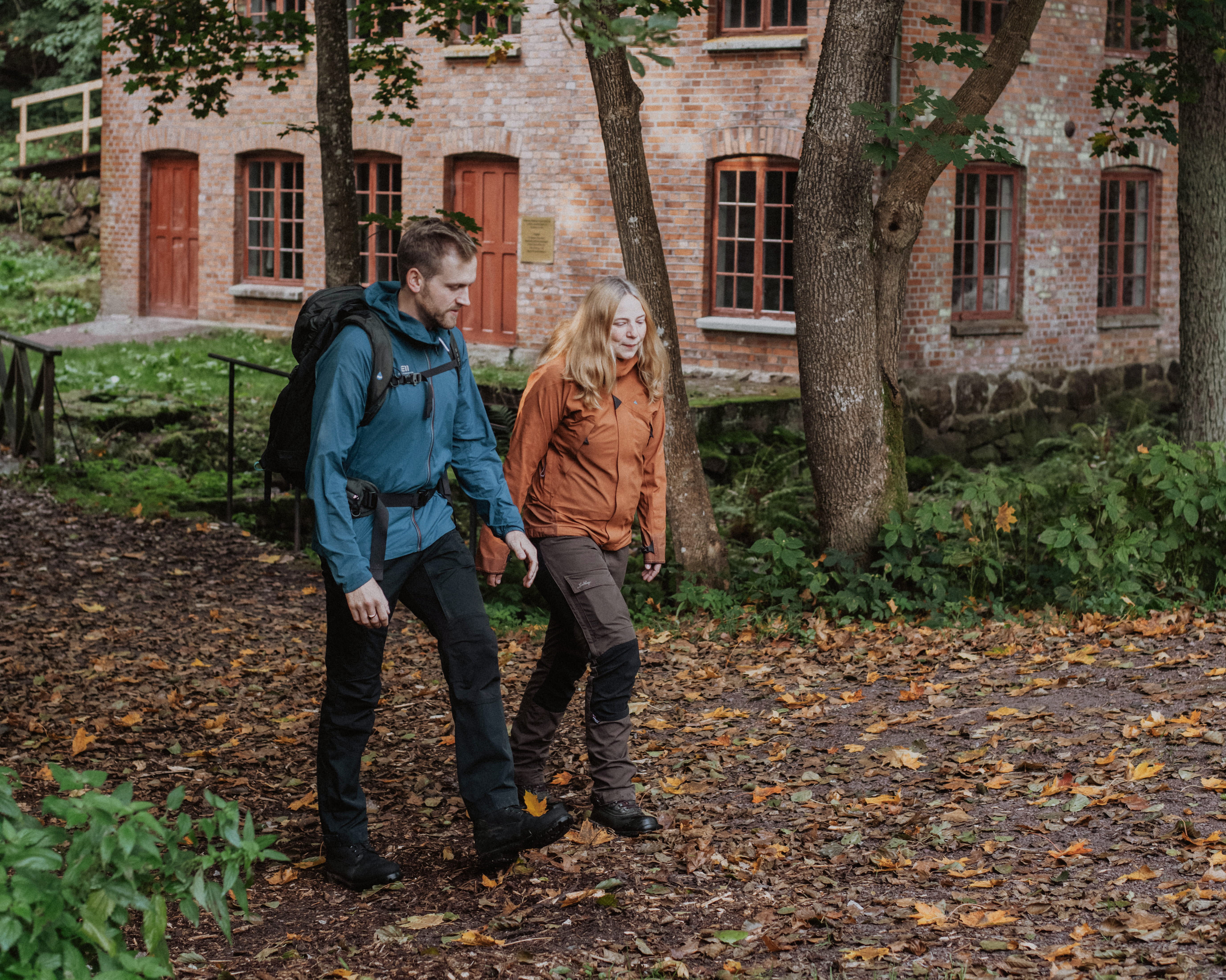 A guy with a blue jacket and a woman in an orange jacket walk on a path by an old brick building.