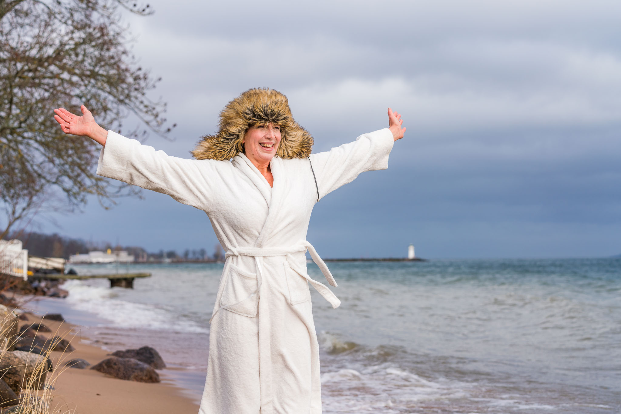 Kvinna står på strand i Hjo mitt i vintern, i morgonrock och pälsmössa. 