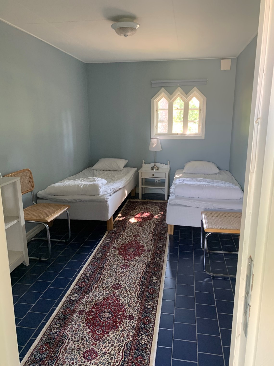 Sovrum med två enkelsängar, ett fönster i mitten och en lång smal matta som ligger på ett mörkgrått klinker golv. 
