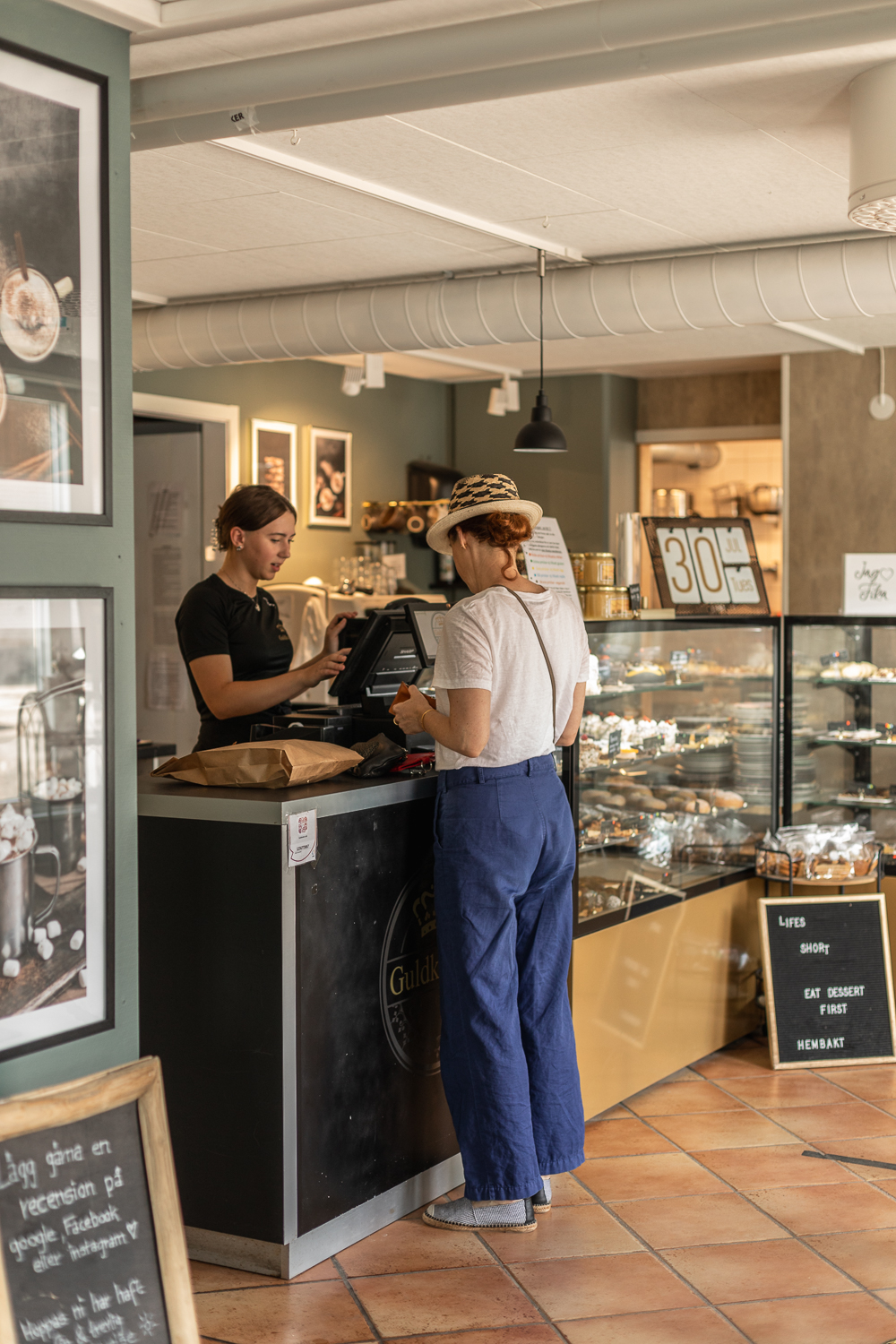En kvinna i hatt står vid en disk på ett café och köper fika av en expedit. 