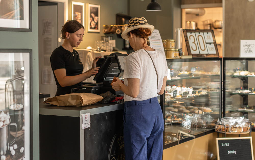 En kvinna i hatt står vid en disk på ett café och köper fika av en expedit. 