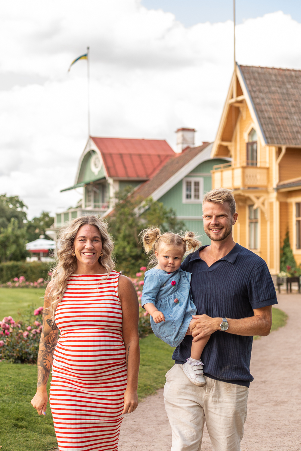 En gravid kvinna i randig klänning går i en park med stora trähus bakom. Bredvid går en man och håller i en liten flicka. 