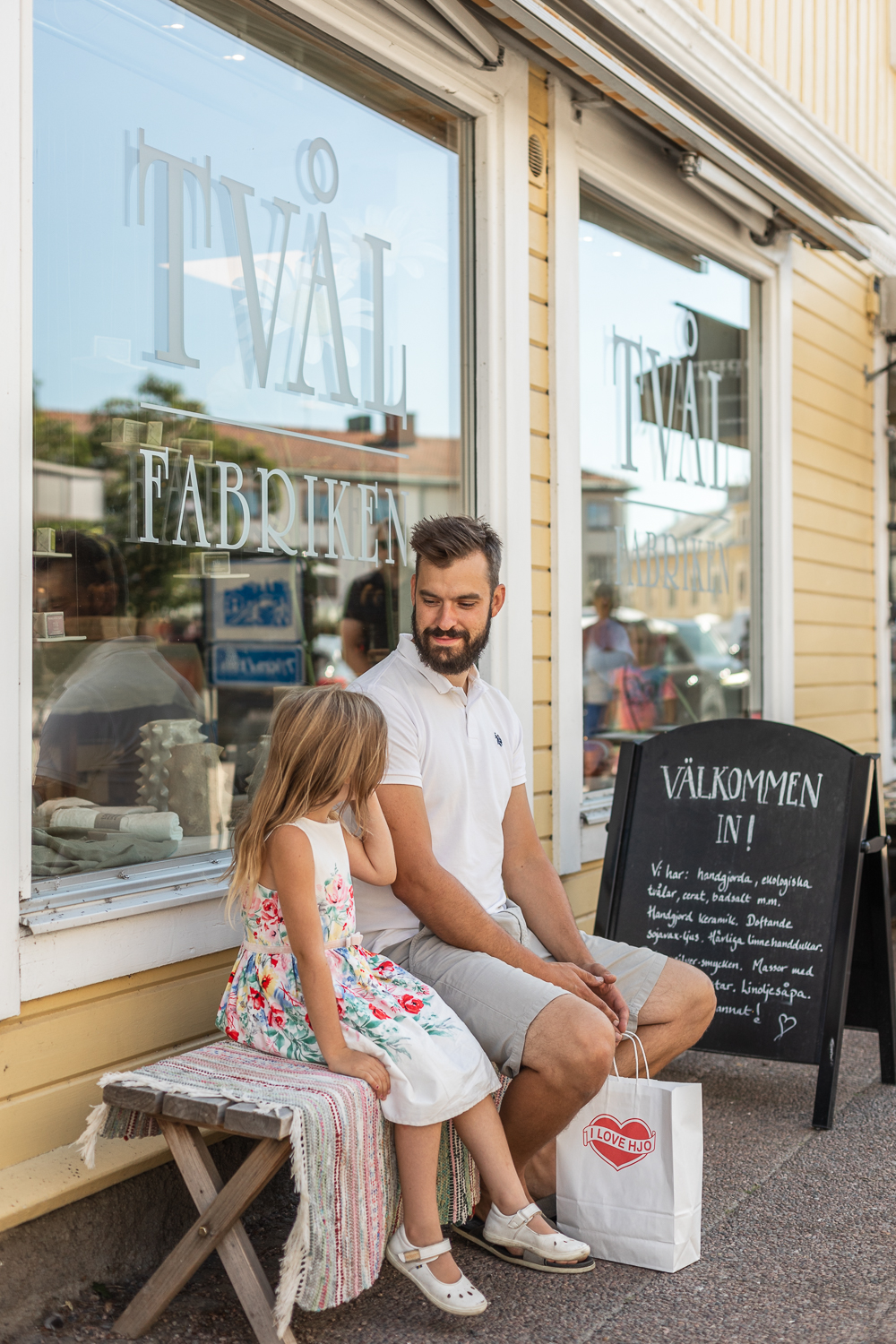 En man sitter ner på en bänk utanför en Tvålfabriken i Hjo med sin dotter. I handen håller han i en kasse där det står I Love Hjo. 