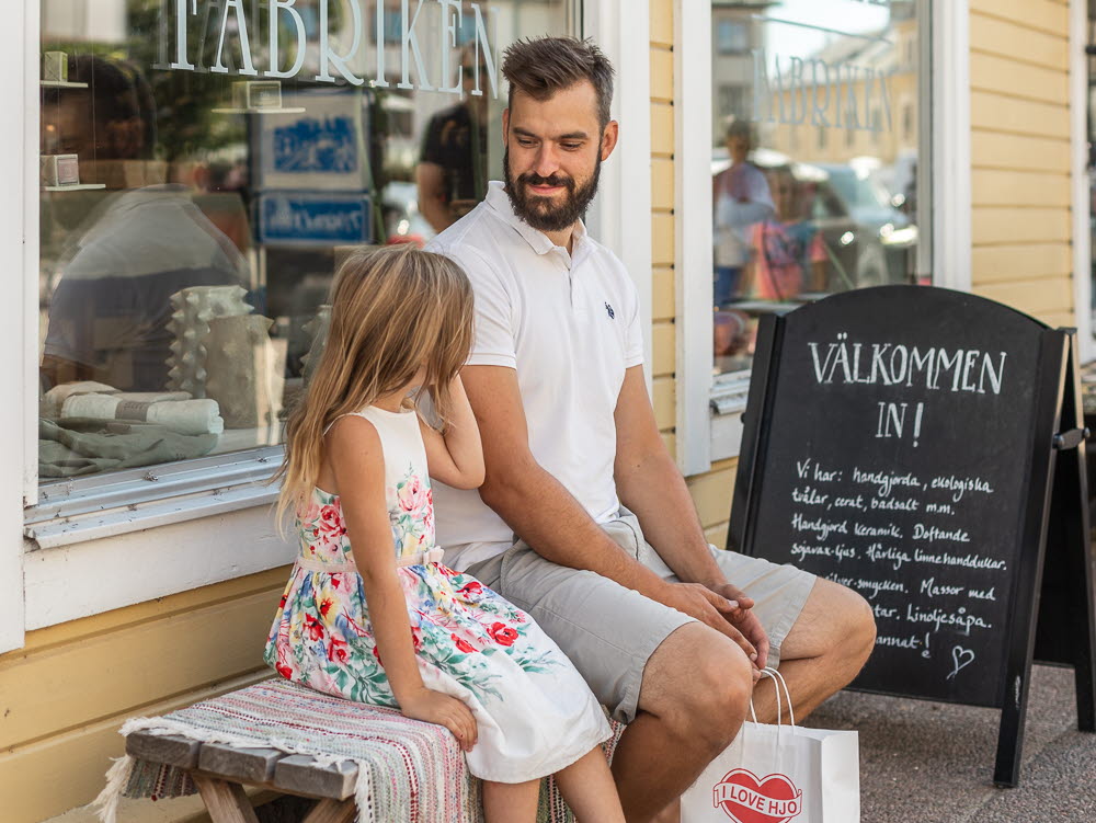 En man sitter ner på en bänk utanför en Tvålfabriken i Hjo med sin dotter. I handen håller han i en kasse där det står I Love Hjo. 