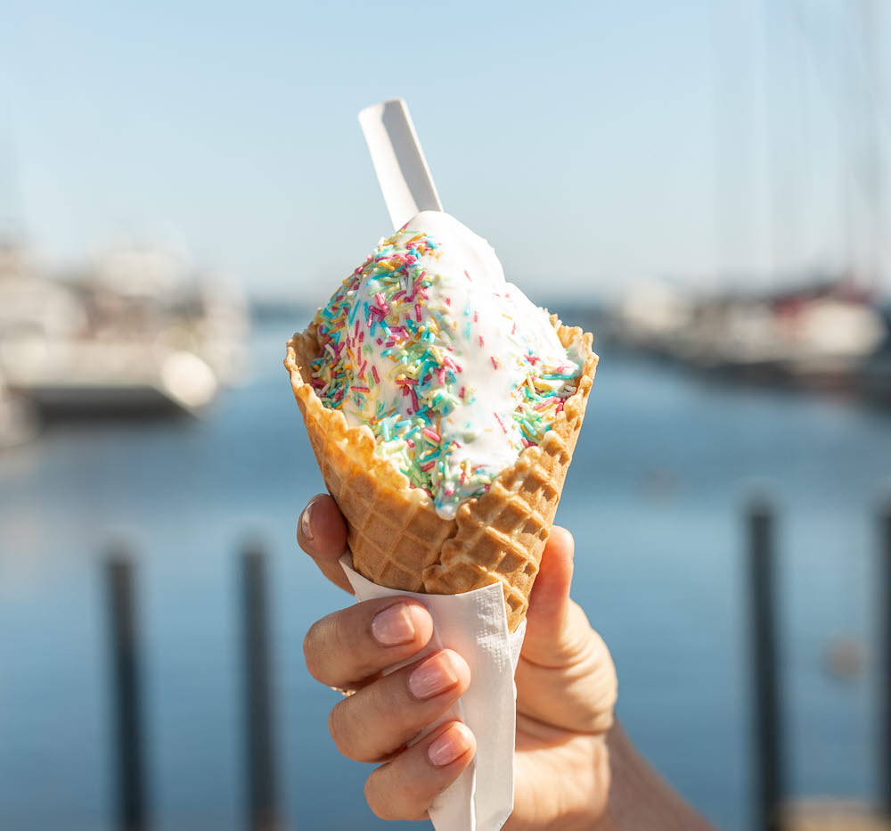 EN hand hålleri en mjukglass i ett rån med strössel på. Bakom skymtar vatten. 