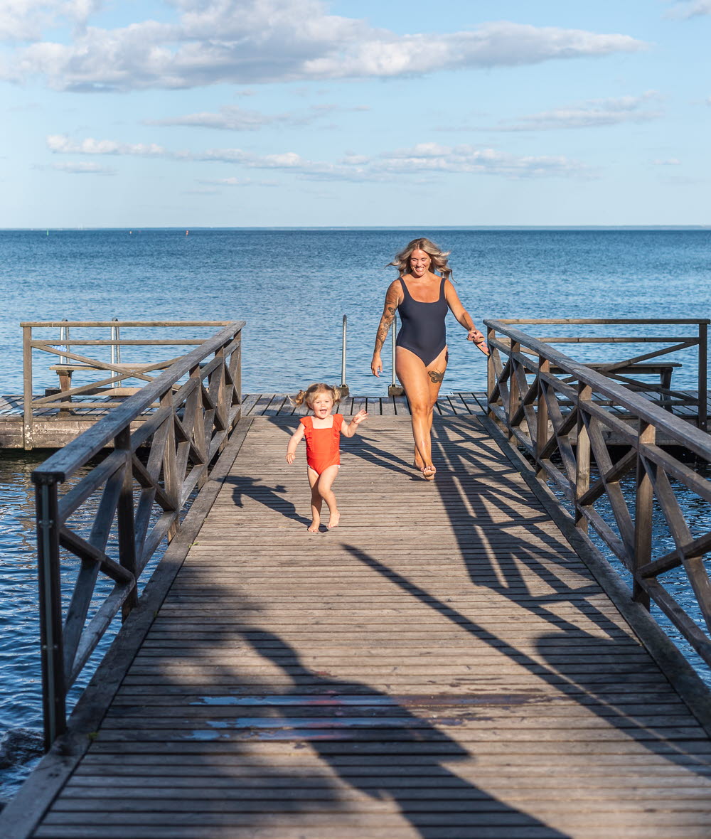 En kvinna i baddräkt går på en brygga och framför henne springer hennes dotter. 